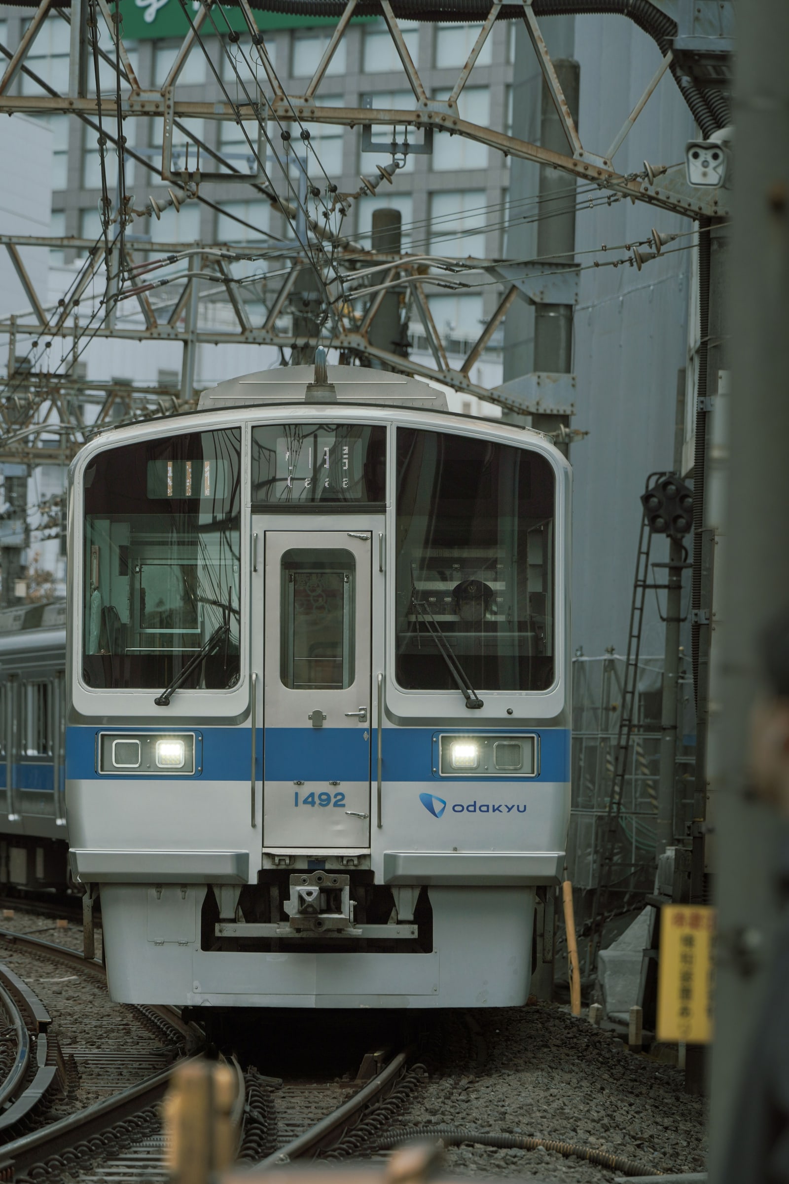 A silver Odakyu commuter train with a blue stripe at a platform under overhead wires and a steel frame.