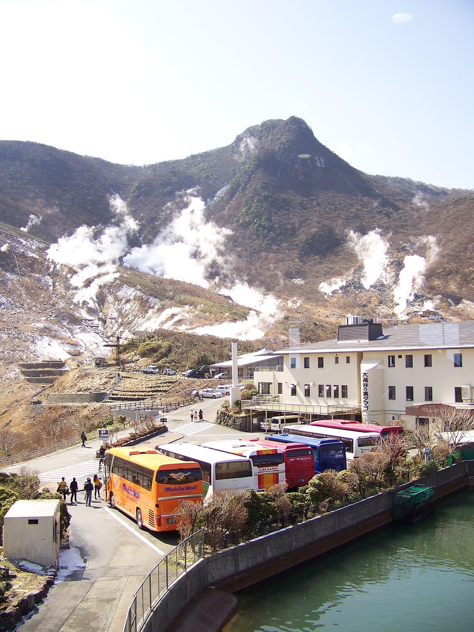 Tour coaches in a row in a parking lot below a steaming volcanic slope and a rocky peak, a white building beside them and people walking on the road.