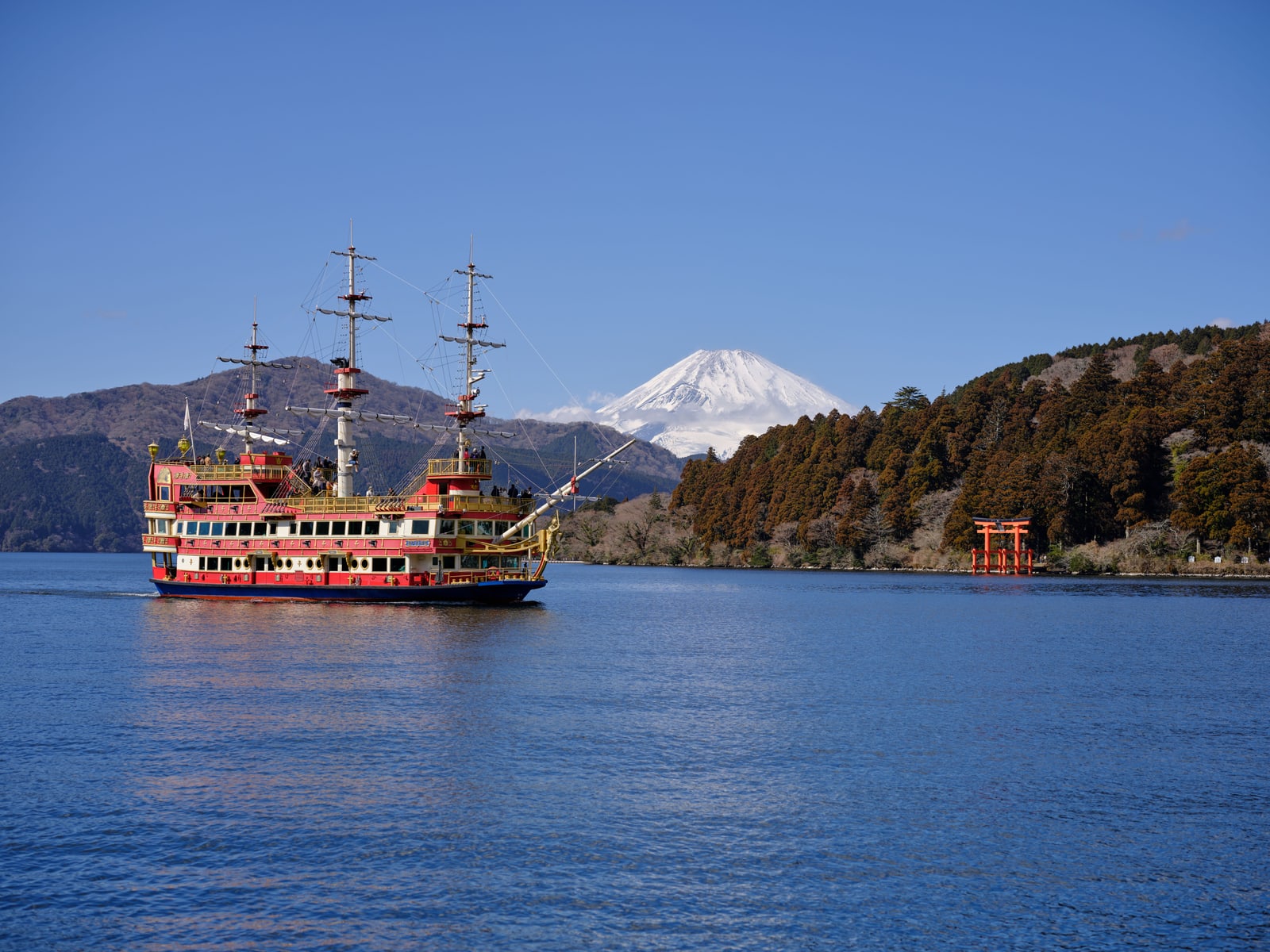 A red and gold sightseeing ship built like a sailing ship crossing Lake Ashi, snow-capped Mount Fuji behind the hills and a red torii on the right shore, under a clear blue sky.