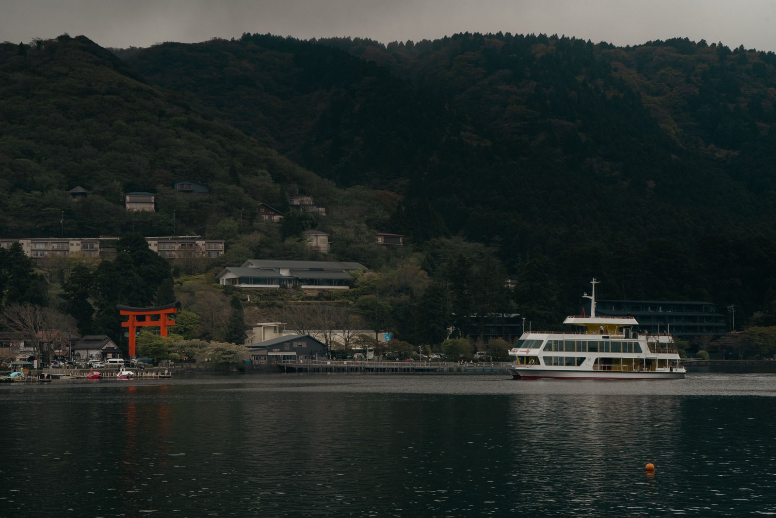 A red torii on the shore of a dark lake below forested hills, buildings among the trees and a white sightseeing boat at a pier.