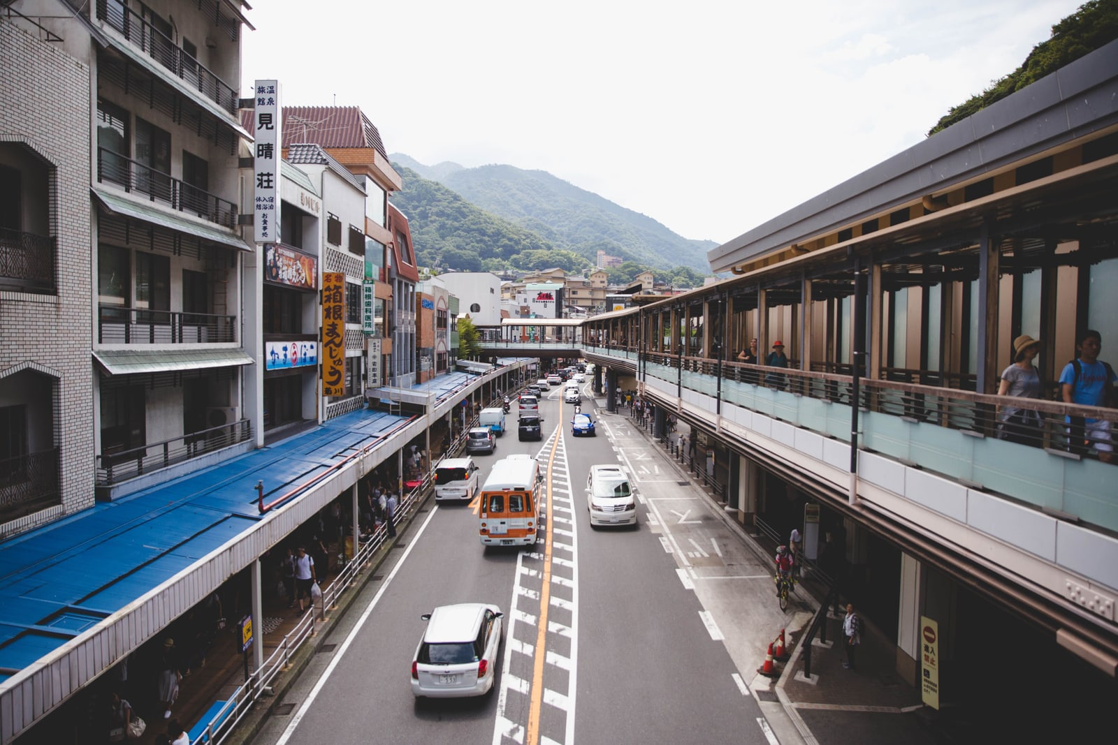 A busy street in a mountain town seen from above, cars and a small bus in both lanes, shops on the left and a covered walkway on the right, green hills behind.