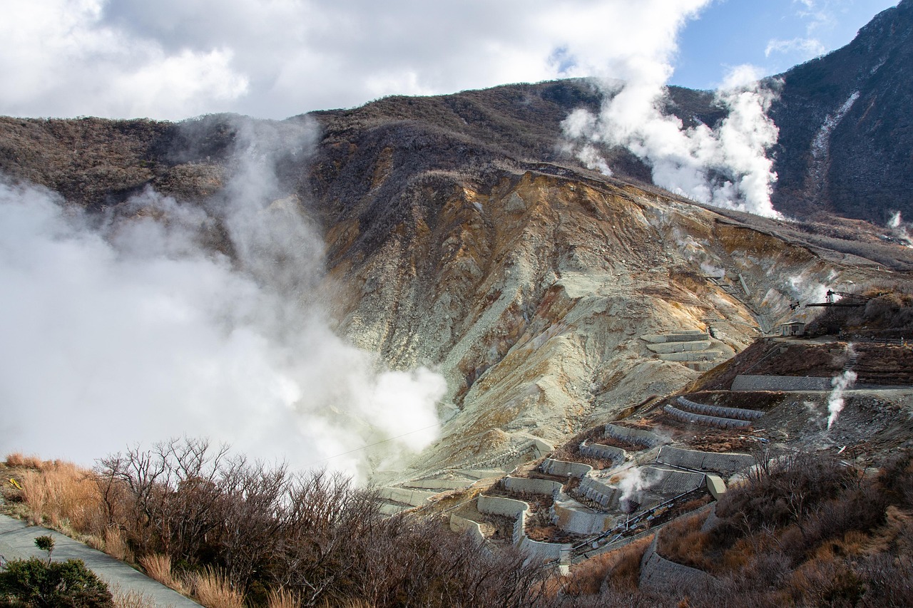White steam pouring from a bare, sulfur-stained volcanic slope, terraced barriers below and dry grass in the foreground.