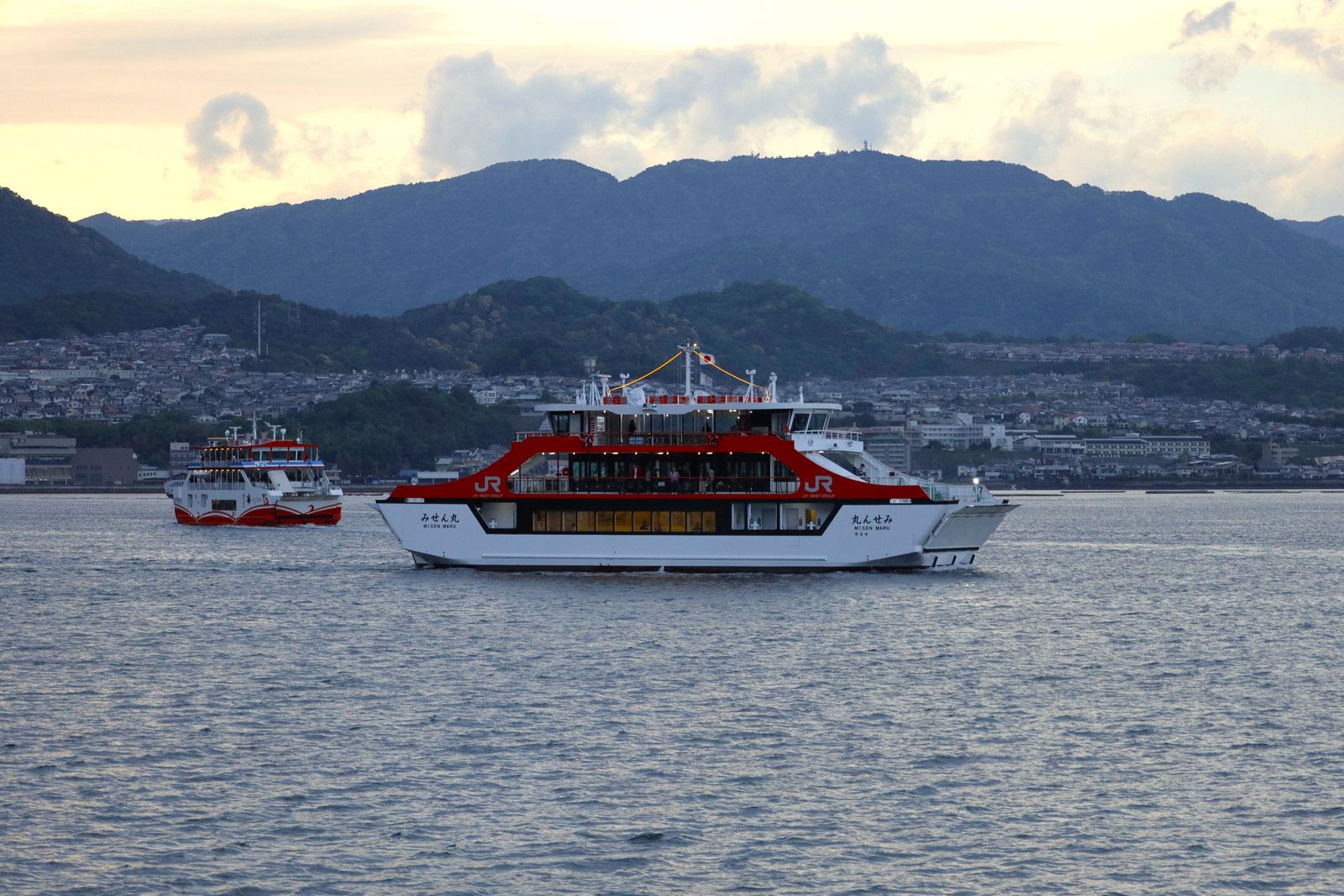 Two red-and-white ferries marked JR crossing calm water, a waterfront town and wooded hills behind in evening light.