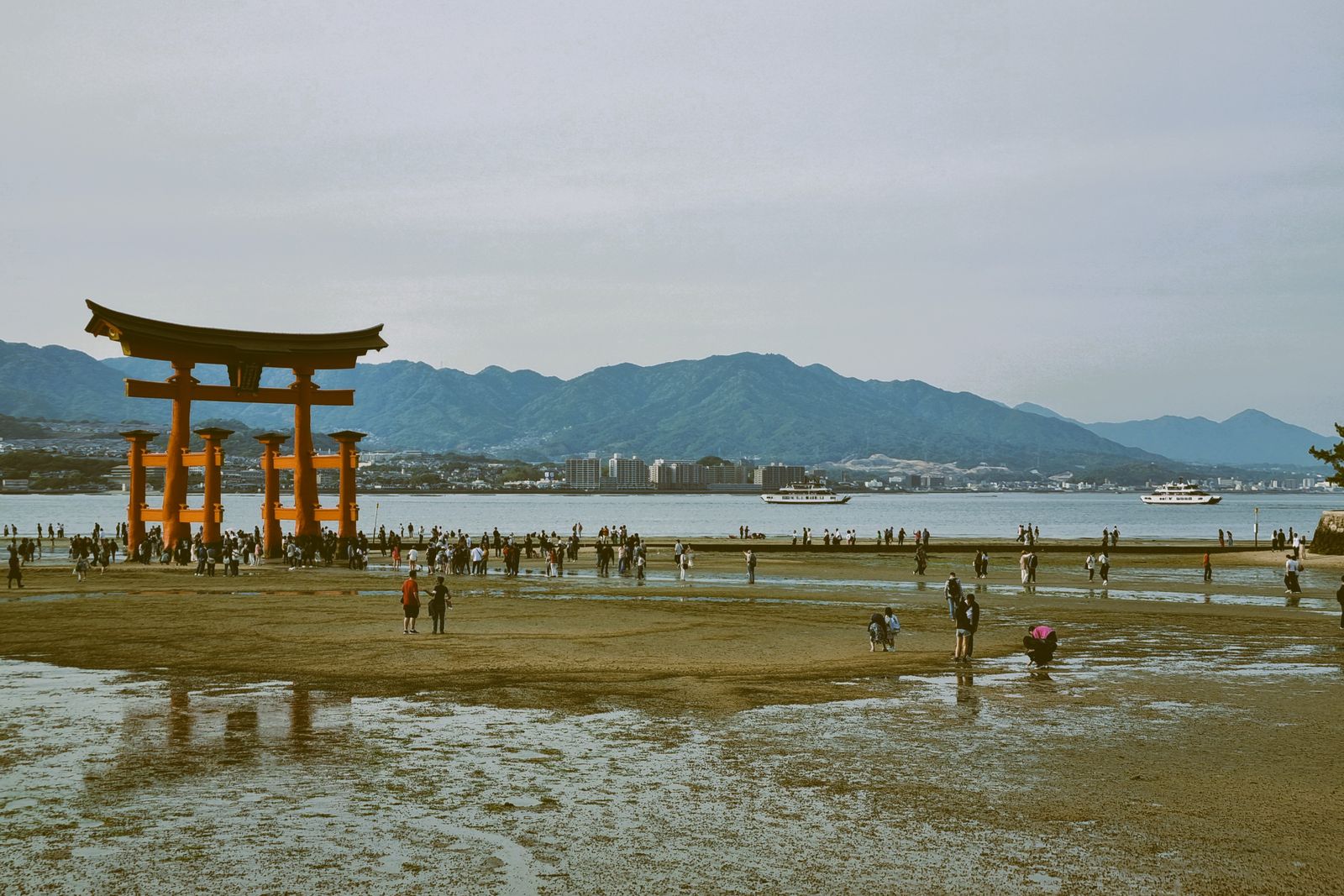 The great torii on an exposed wet tidal flat, dozens of small figures walking around its base under a grey sky.