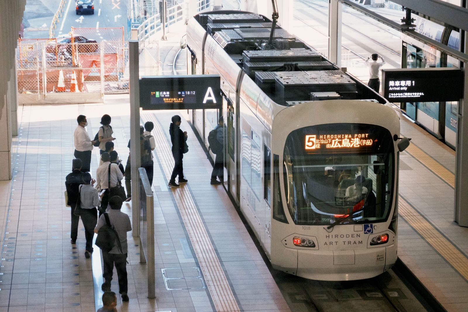 A white Hiroden streetcar at a covered platform at Hiroshima Station, passengers waiting on the platform.