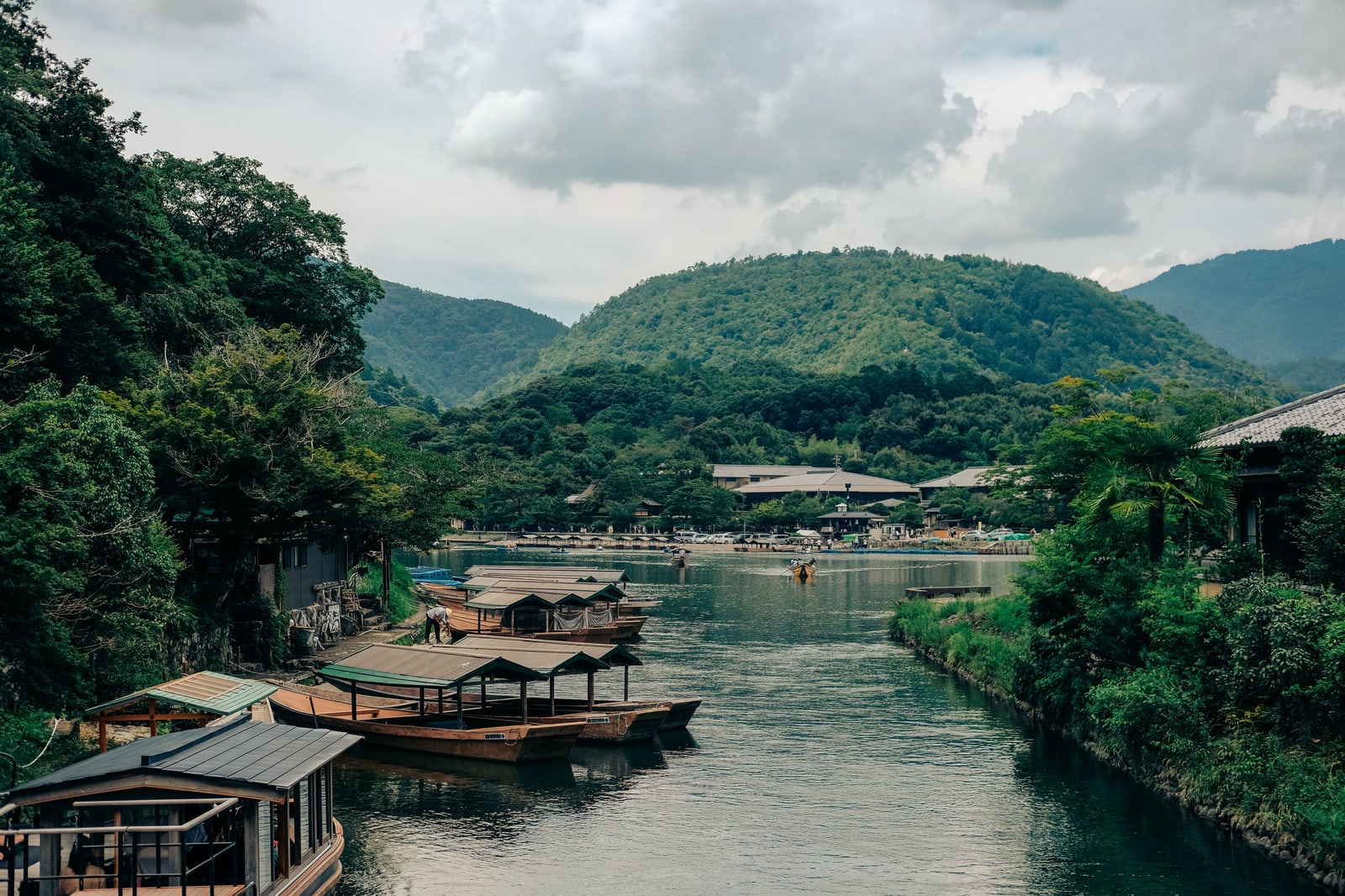 Roofed wooden river boats moored along the riverbank in Arashiyama, green hills in the distance.