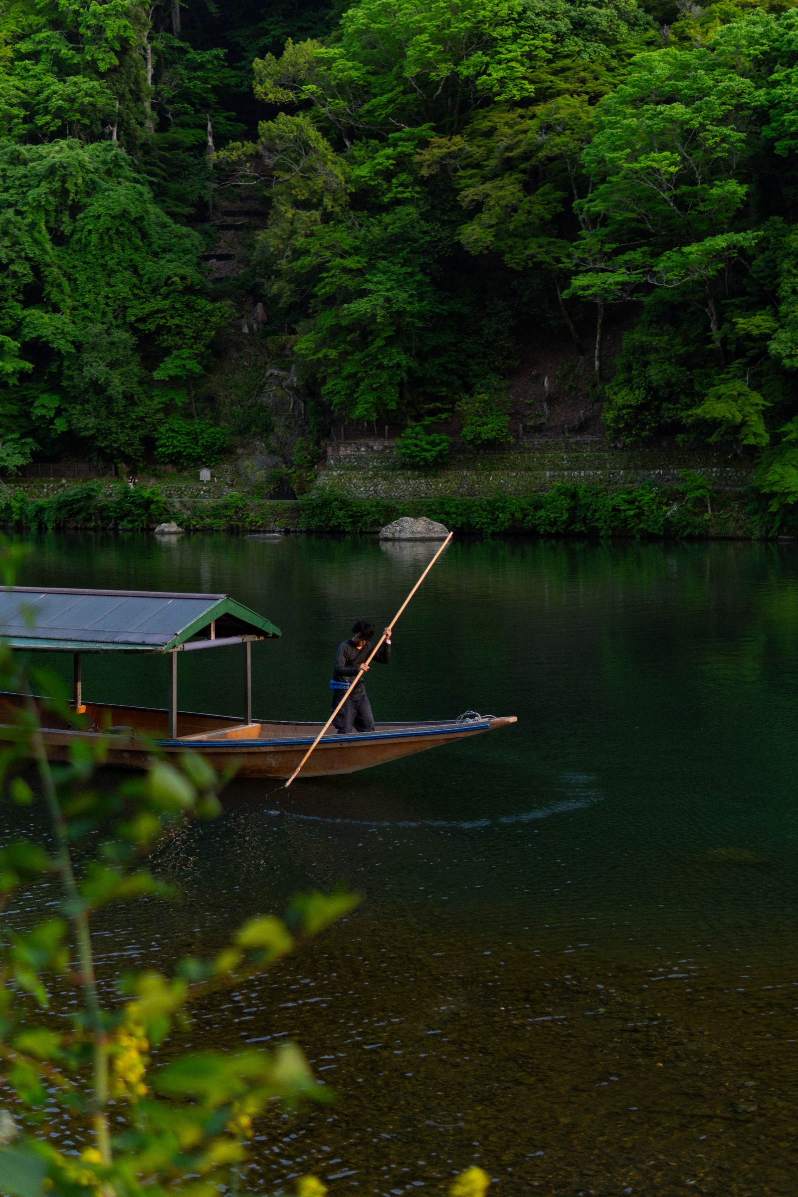 A small roofed wooden boat poled along a dark green river at the foot of a forested slope.