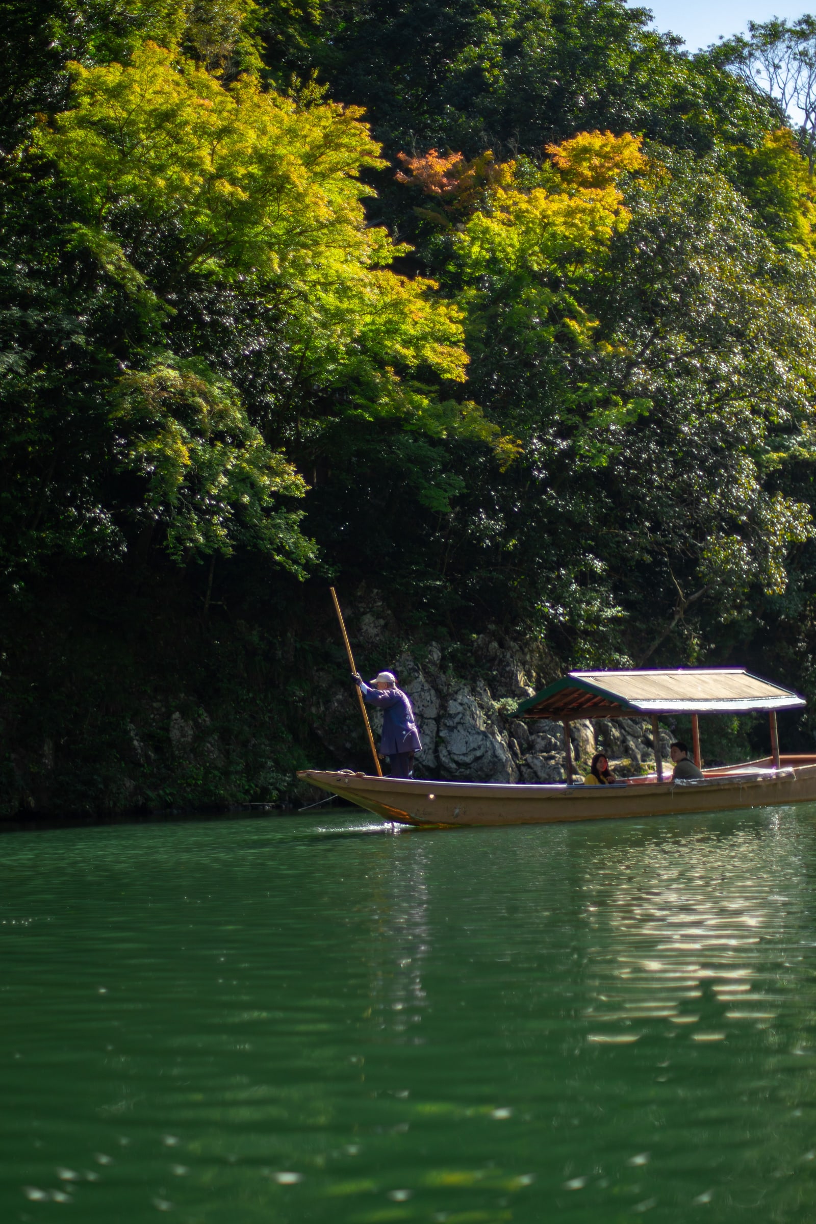 A long wooden boat with a canopy poled along deep green water under a wooded rock bank, a boatman standing at the bow.