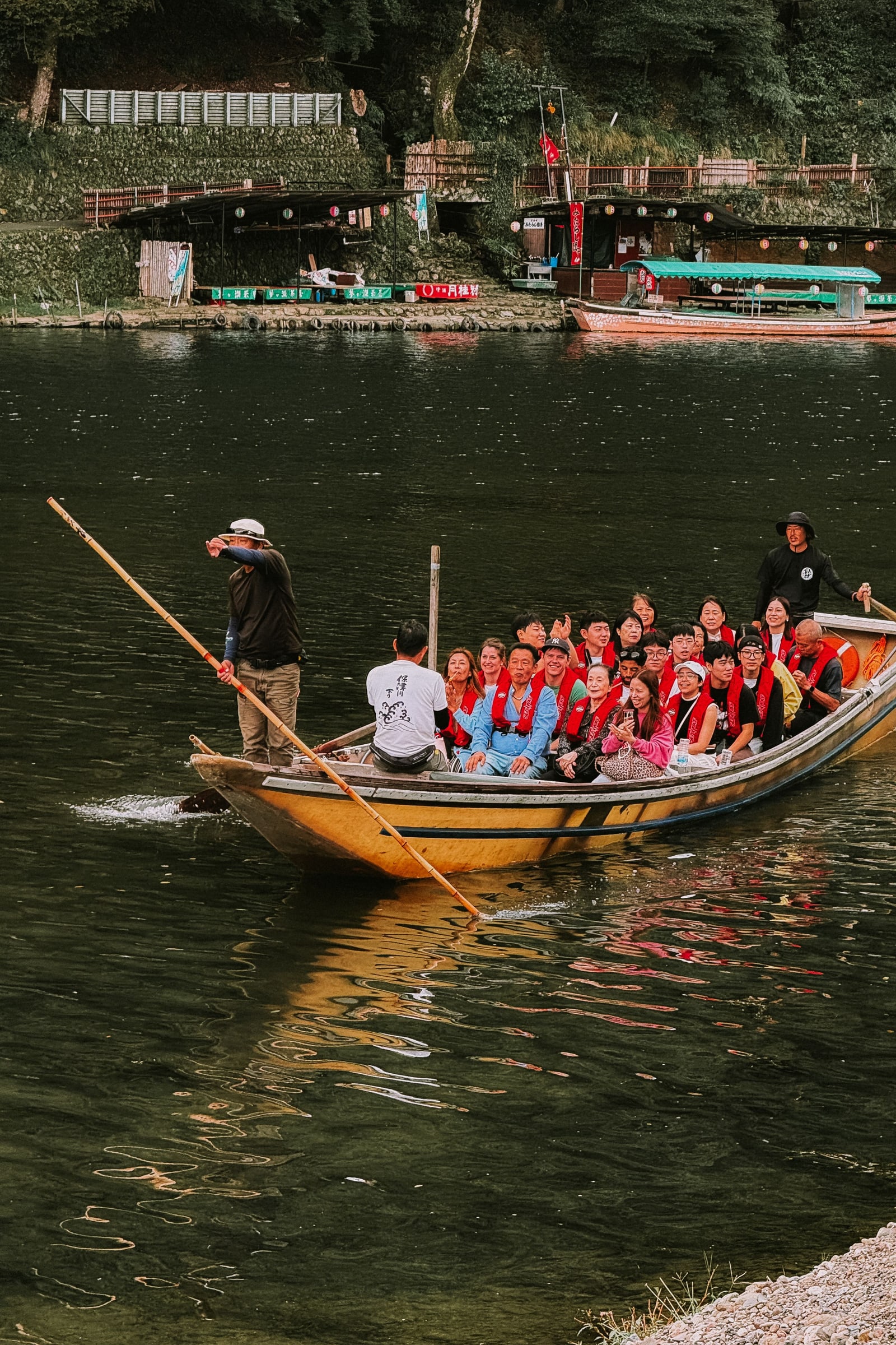 A long yellow wooden boat full of passengers in red life jackets, one boatman poling at the bow and another at the stern, riverside teahouses behind.