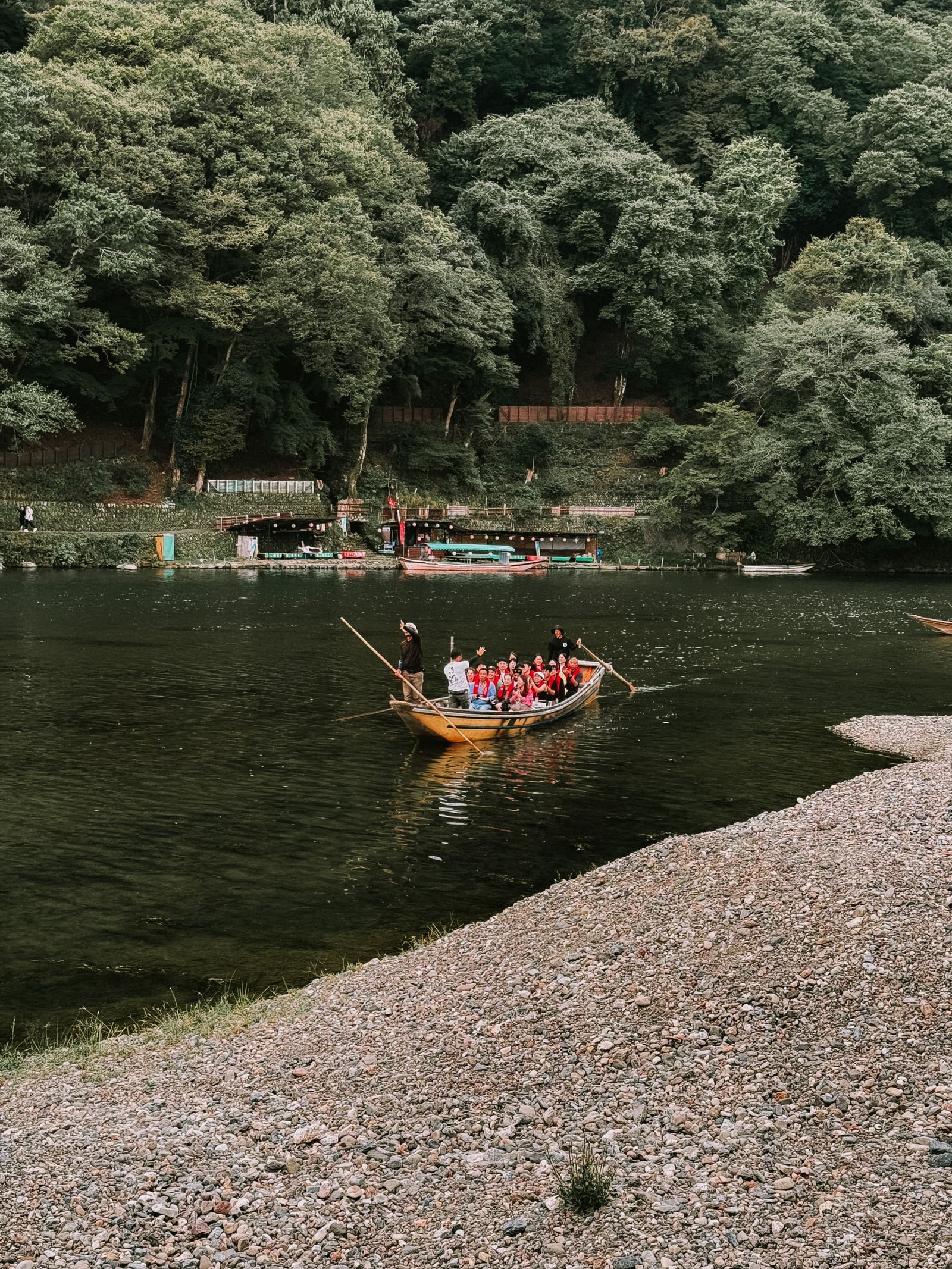 A wooden boat of passengers in red life jackets passing a wide gravel bar below a wooded hillside.
