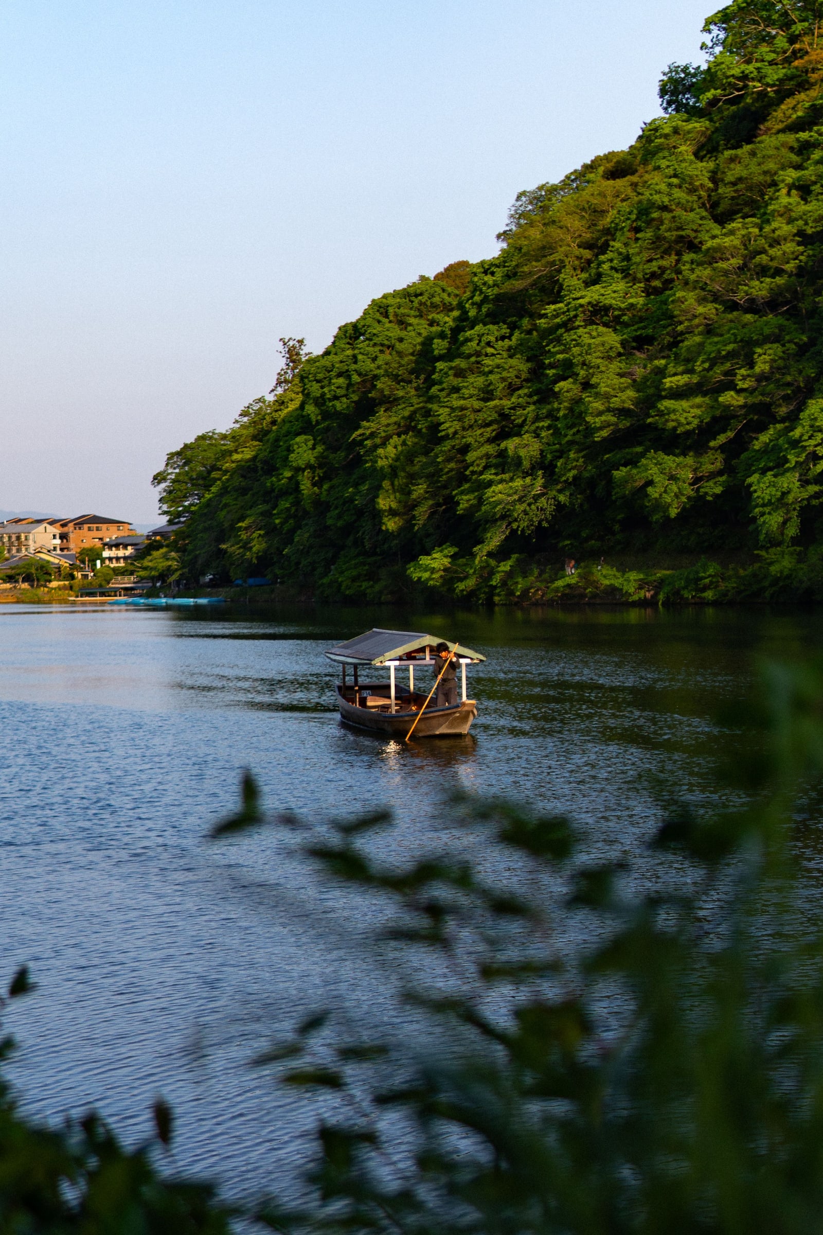 A small roofed boat on a wide, calm river below a wooded hill, low houses on the far bank in evening light.