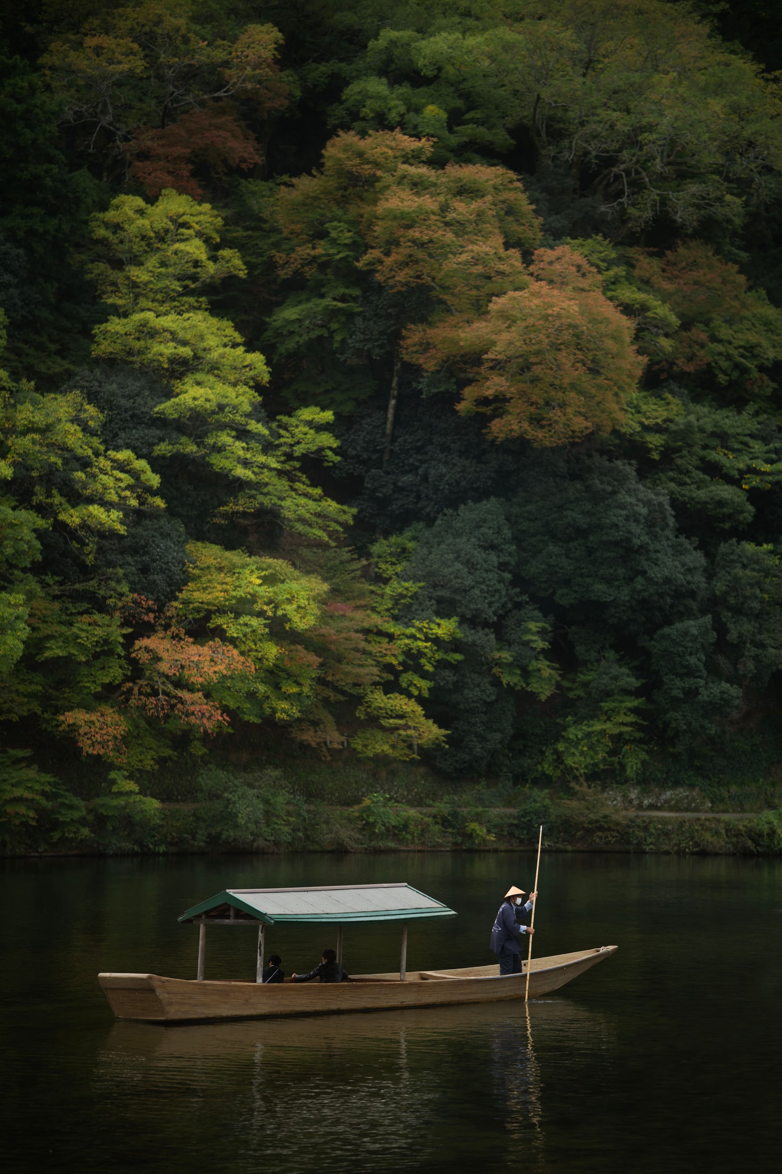 A roofed wooden boat on a calm river, a boatman with a long pole at the stern, early autumn color on the hillside behind.