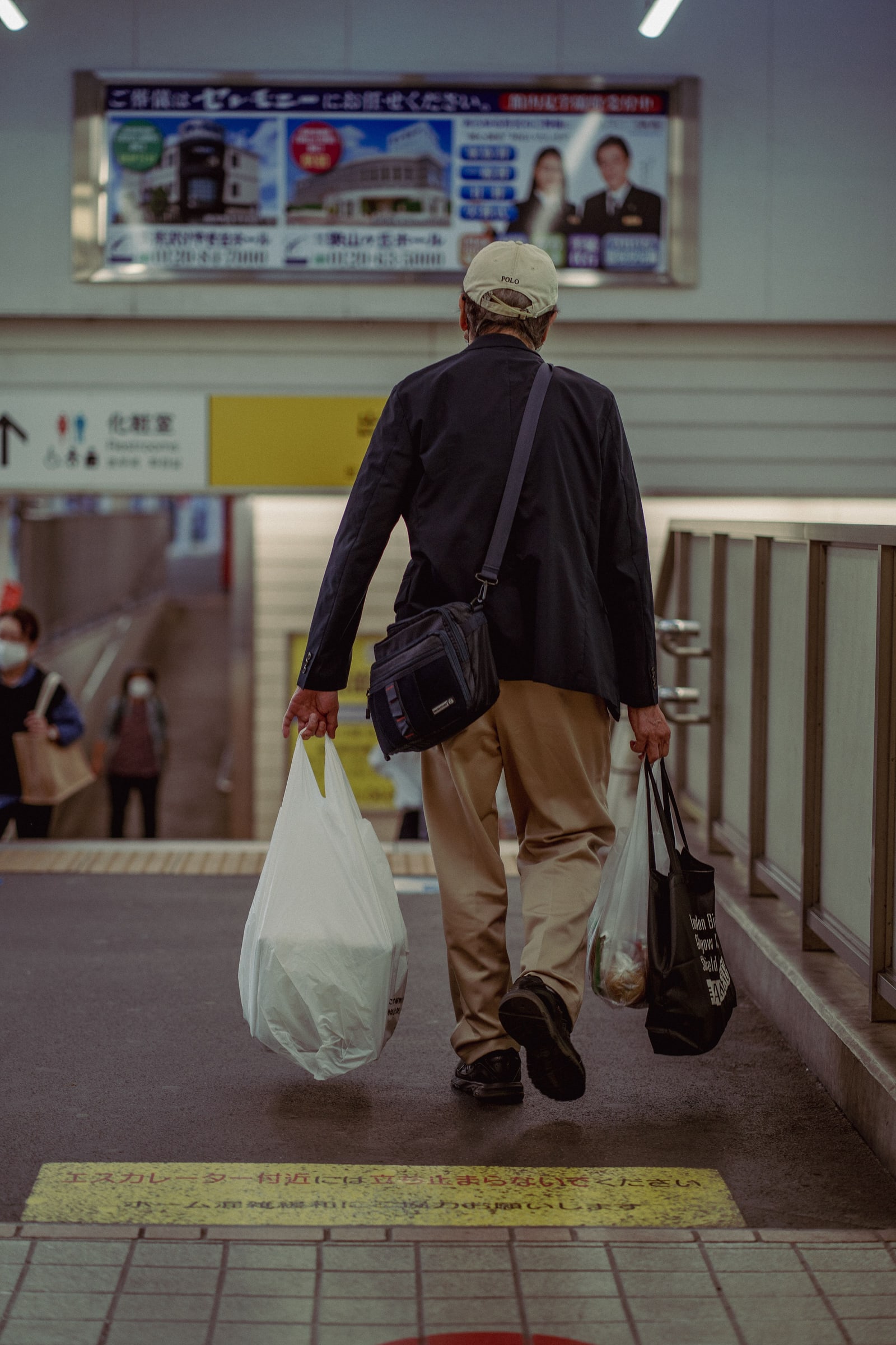 A man seen from behind walking through a station ticket gate with a shopping bag in each hand.