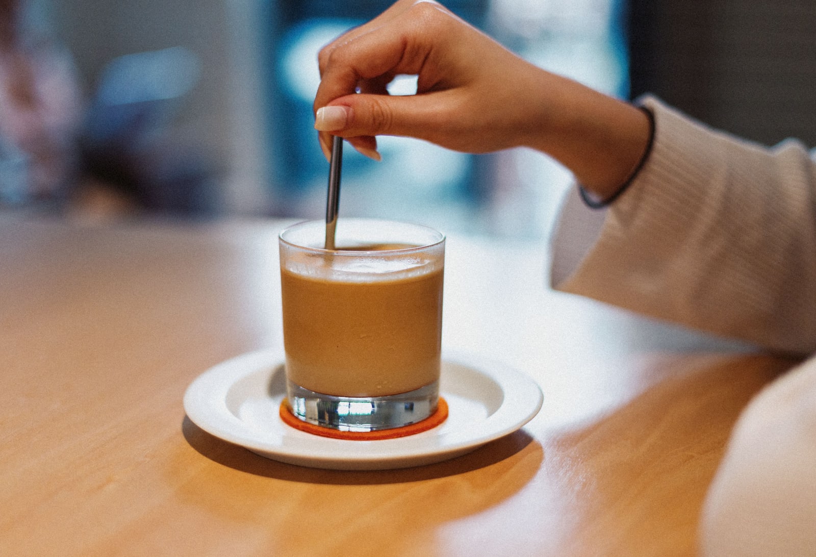 A hand stirring a glass of milky coffee on a saucer on a wooden café table.