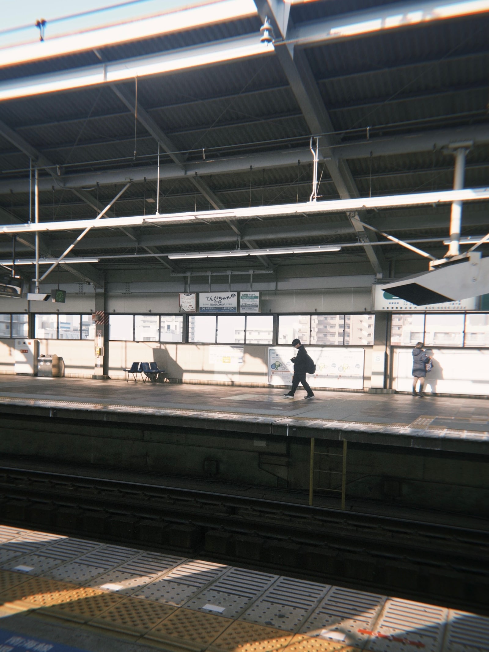 Two people waiting on a sunlit train platform in Japan, the tracks in front of them.