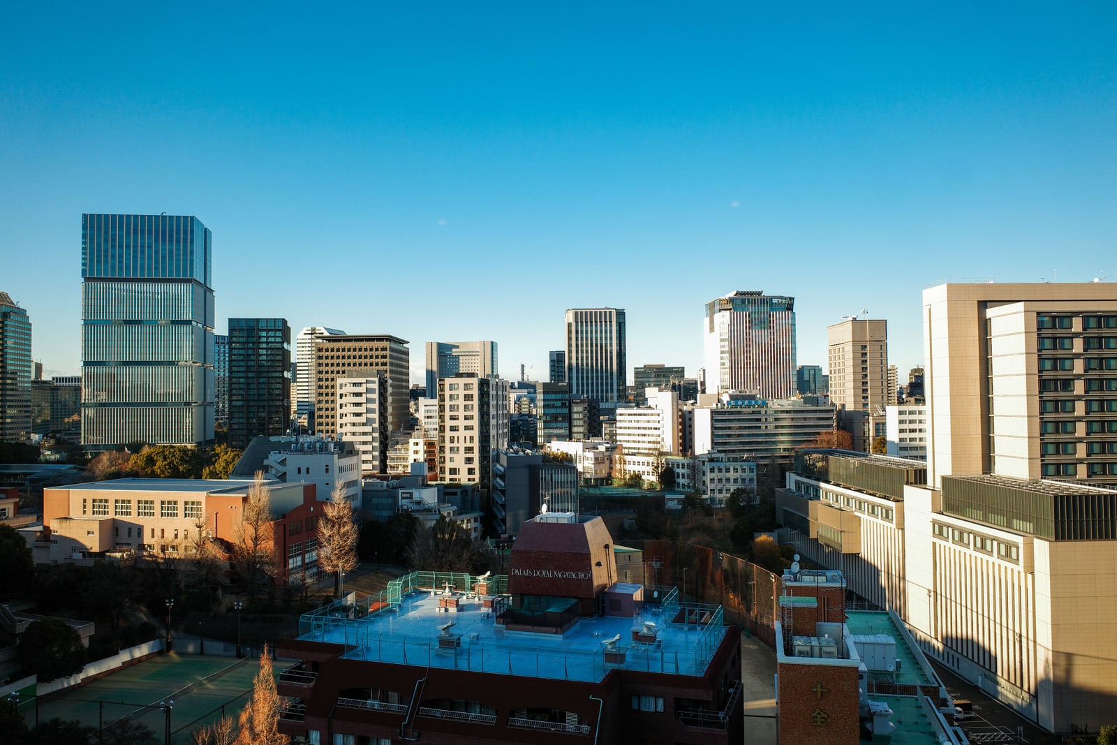 Office towers and lower city blocks of central Tokyo under a clear blue sky, seen from high above.