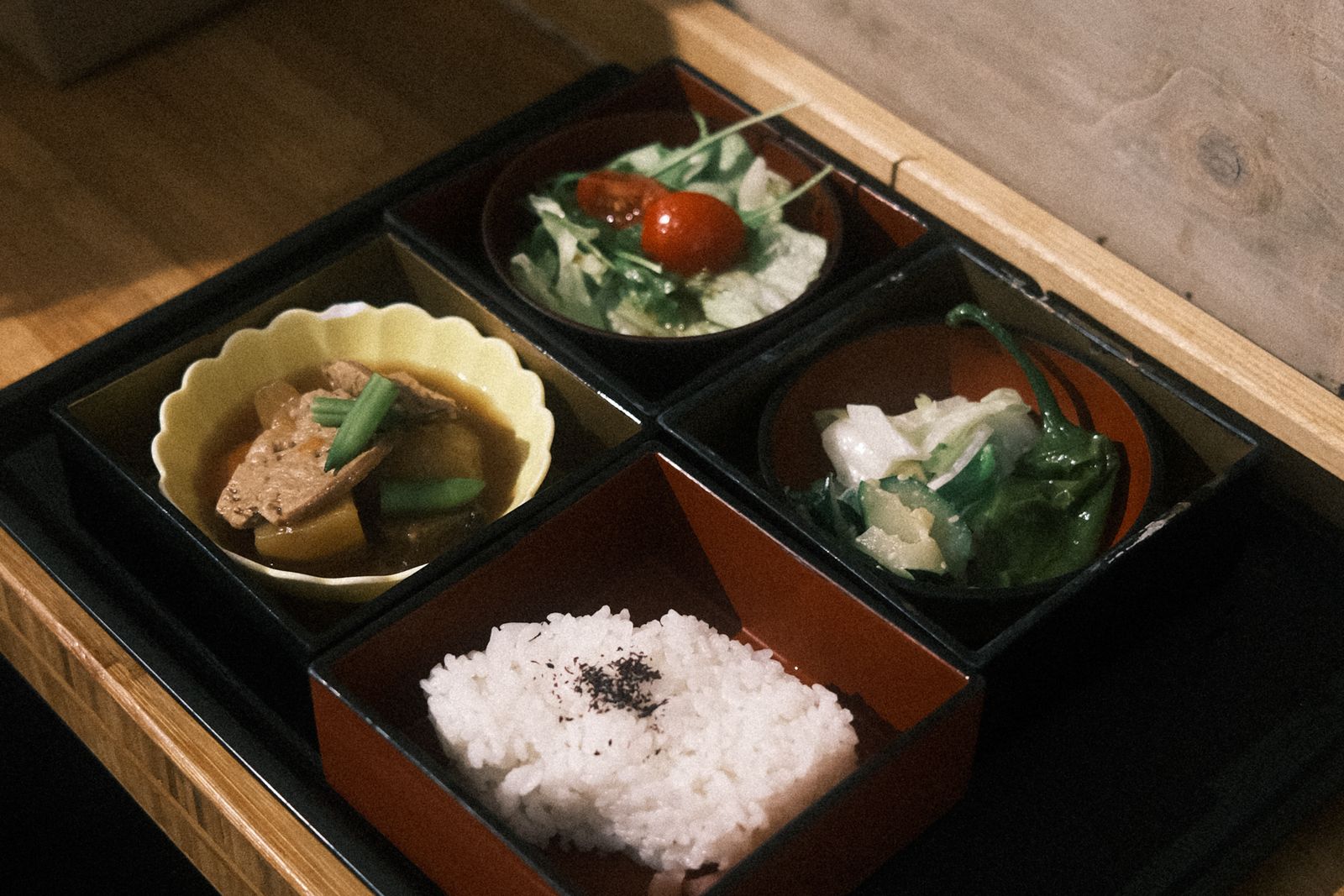 A four-compartment black-and-red lacquer-style box on a wooden tray — rice in one compartment, a simmered dish in a yellow fluted bowl, a salad with cherry tomatoes, a green vegetable.