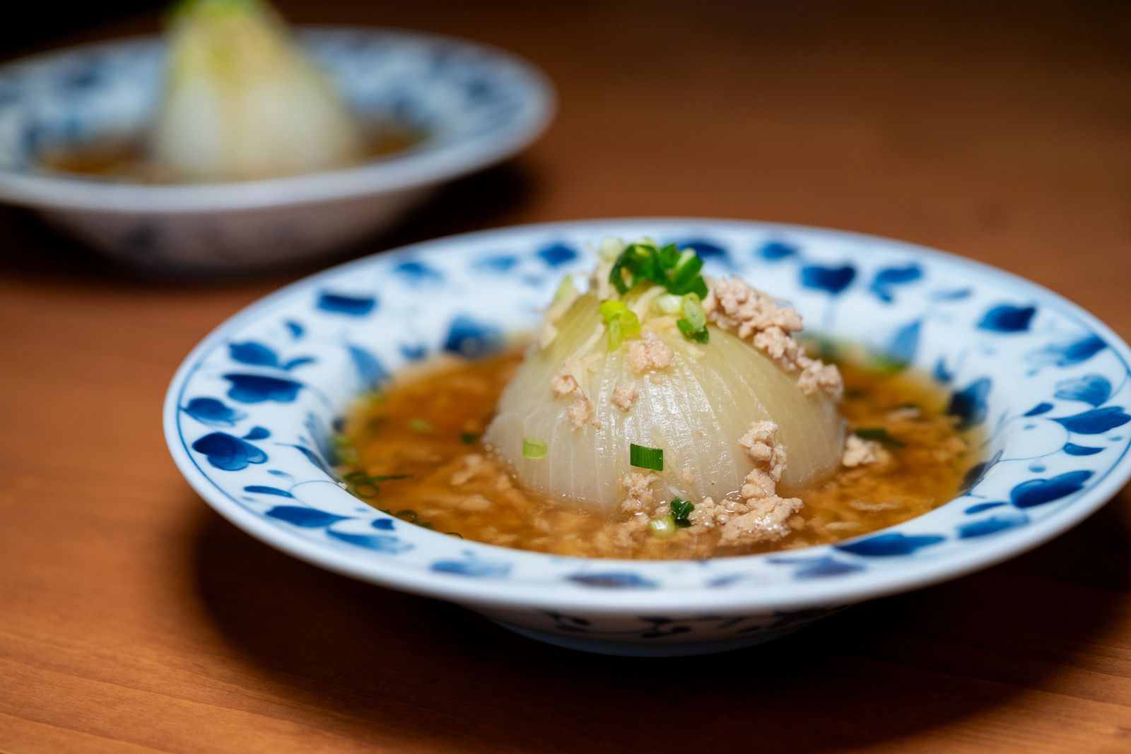 A shallow blue-and-white floral-rimmed dish holding a whole simmered onion in broth, a second dish soft in the background.