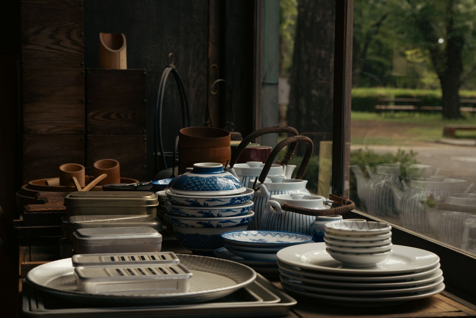Two flat aluminum box graters on metal trays in the foreground, stacks of small dishes, bowls and teapots behind, trees through a window.