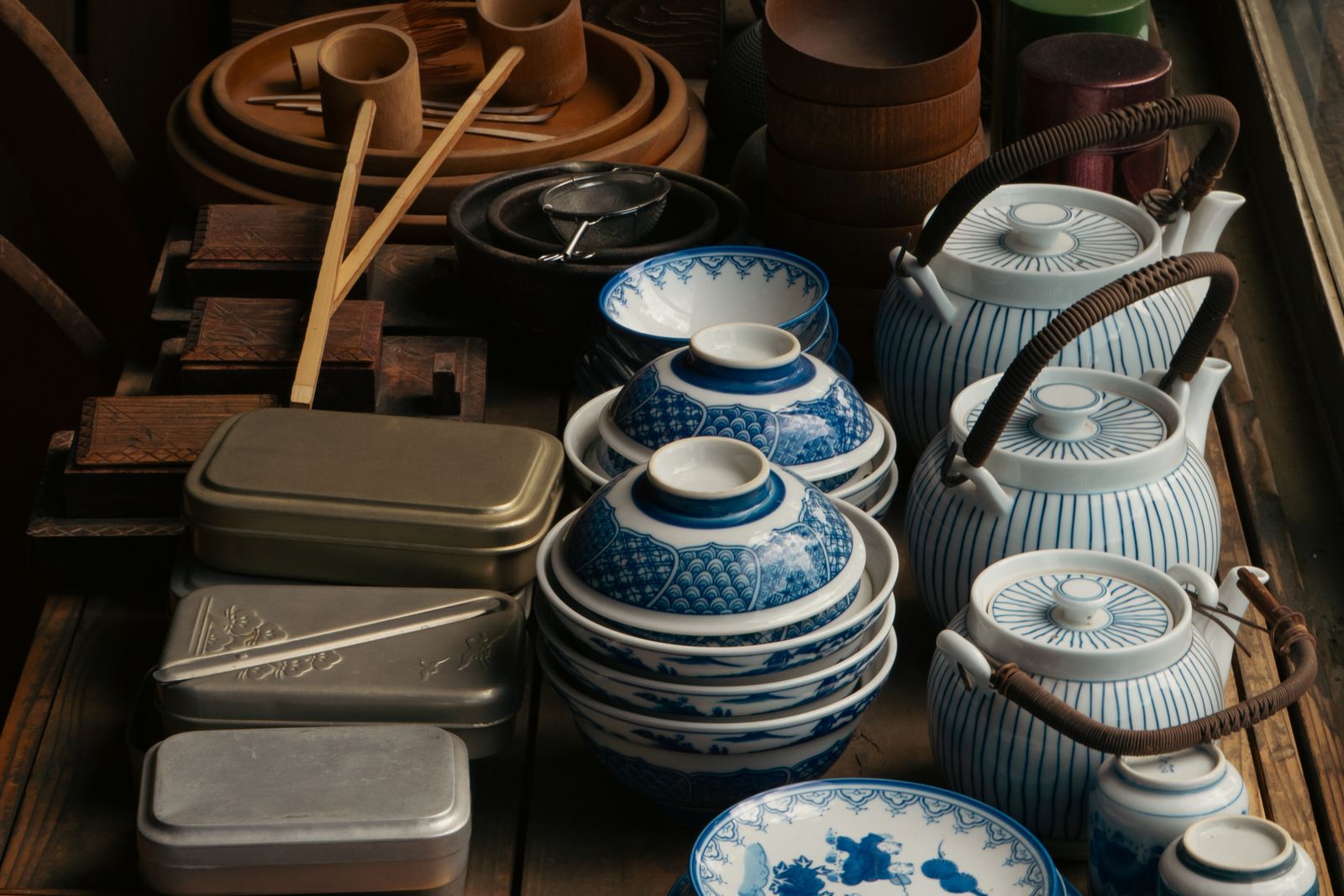 Household tableware on display — stacked blue-and-white rice bowls, lidded bowls, striped porcelain teapots with rattan handles, aluminum lunch boxes and a stack of small plates.