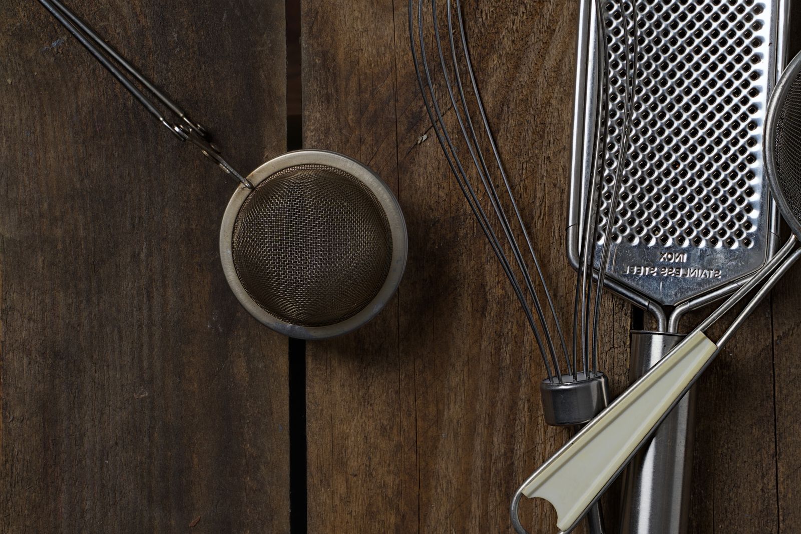 A flat lay of steel kitchen tools on dark planks — a mesh strainer, a whisk, a flat grater and a peeler.