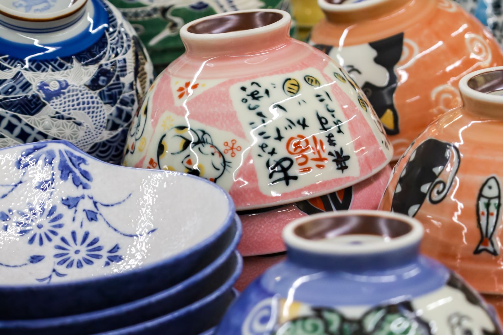 A close, stacked display of patterned porcelain rice bowls — pink with a cat, orange with fish, blue florals, indigo carp.
