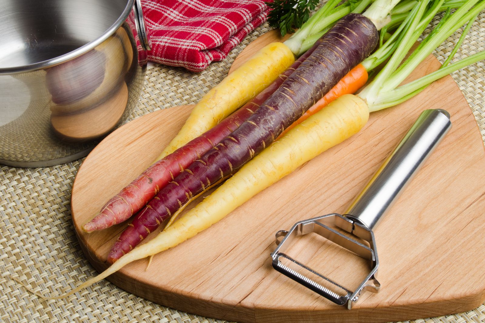 A steel Y-peeler beside purple, yellow and orange carrots on a round wooden board, a steel pot and a red checked cloth behind.