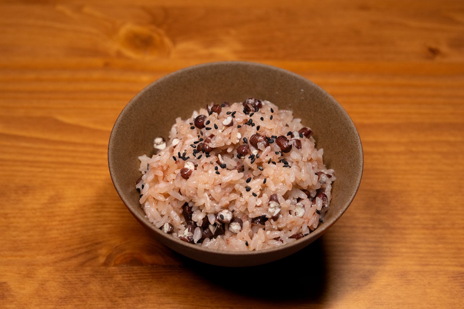 One brown stoneware rice bowl on a pine table, holding rice cooked with red beans and black sesame.
