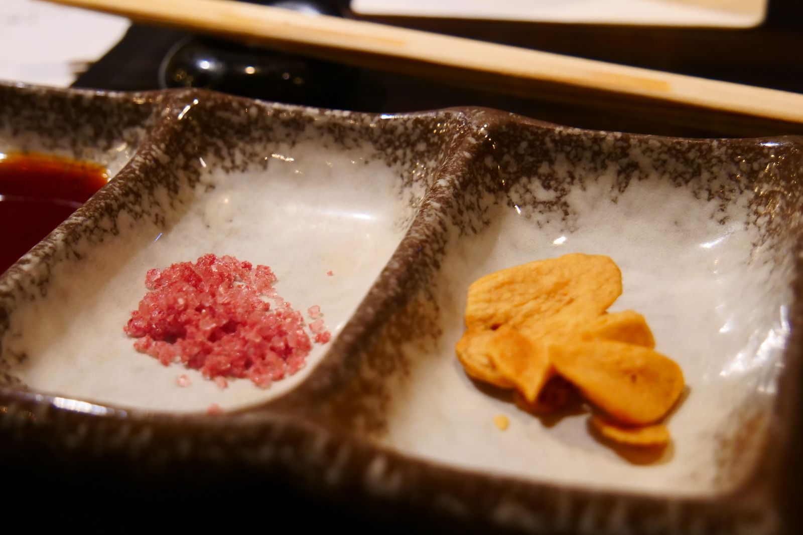A three-compartment speckled stoneware condiment dish holding a dark sauce, pink salt and fried garlic chips, chopsticks on a rest behind.