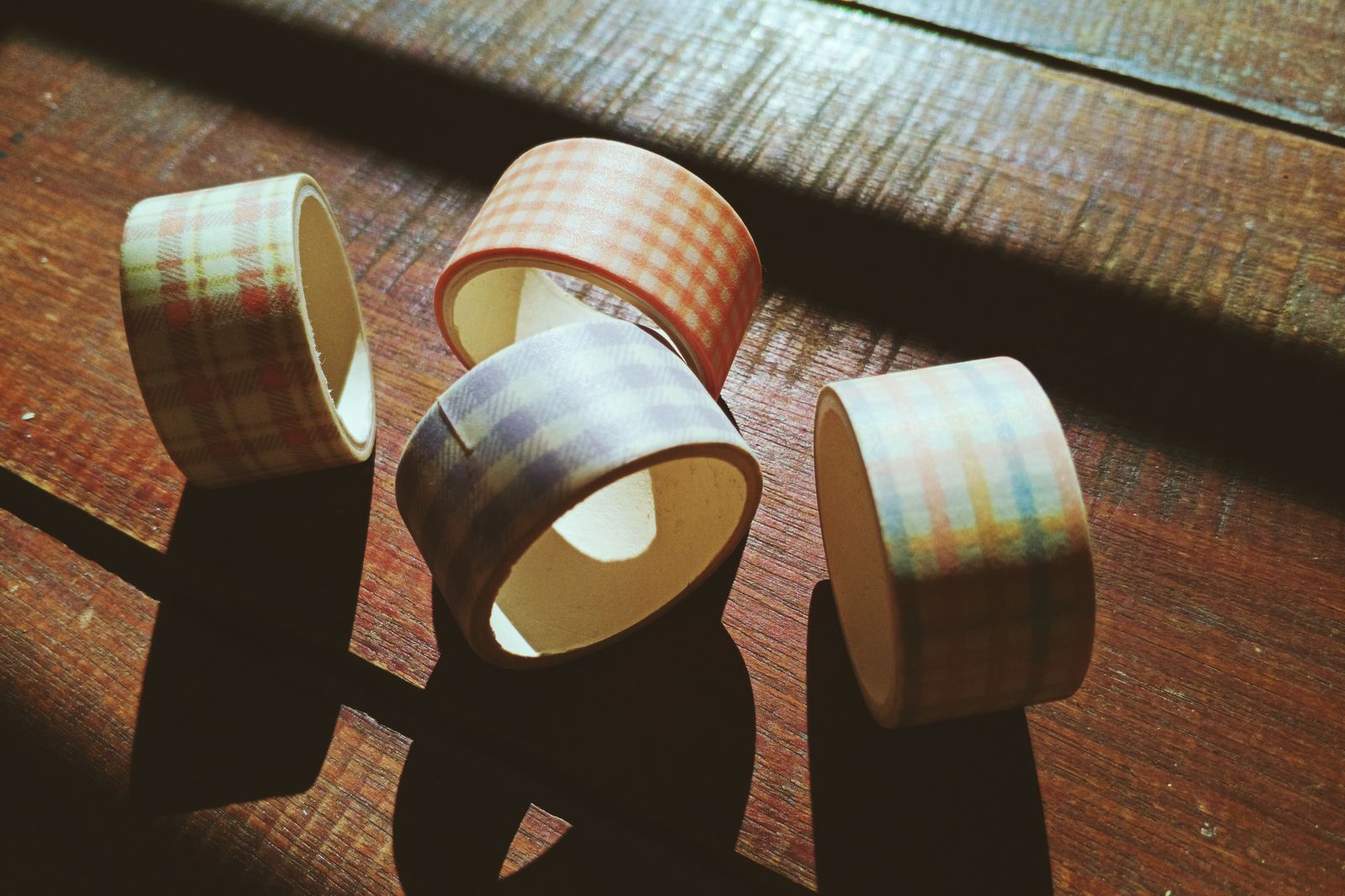 Four rolls of gingham-patterned paper masking tape standing on a sunlit wooden surface, casting long shadows.