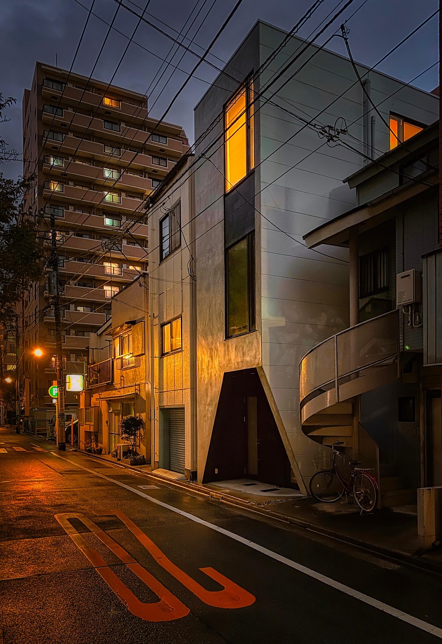 A wet back street at dusk with a pale angular house showing lit orange windows, a spiral stair and a parked bicycle at right, and a mid-rise apartment block behind