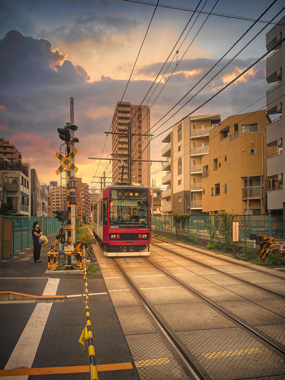 A red-and-white Toden Arakawa Line tram approaching along street-level tracks at a level crossing at sunset, with apartment blocks on both sides and a crossing signal at left