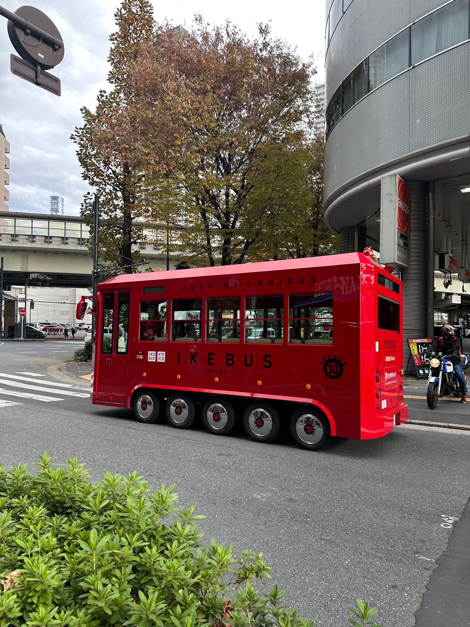 The small red IKEBUS bus of Toshima City at a street junction in autumn, with trees and an elevated road behind it