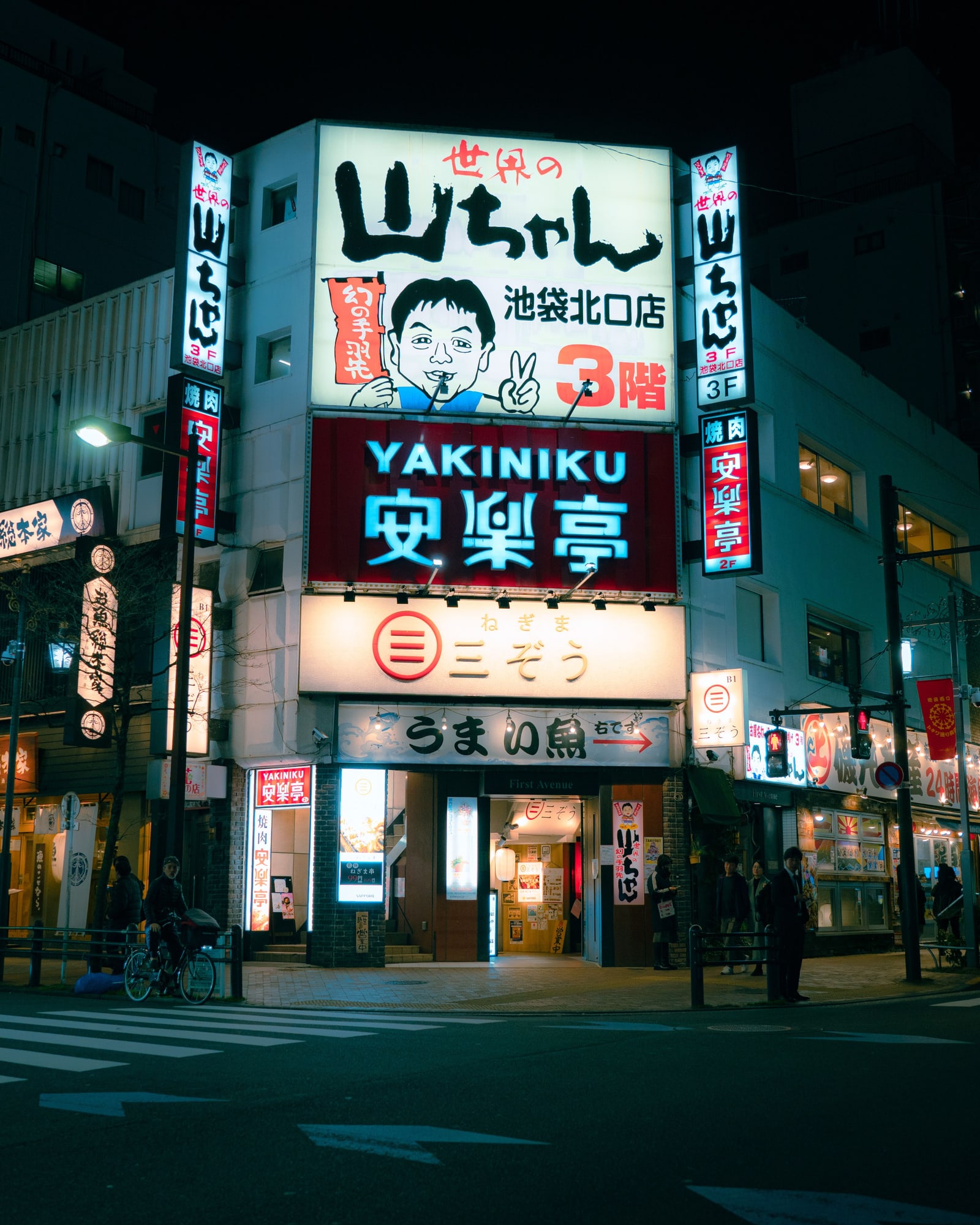 A street corner in Ikebukuro at night, a building covered in lit restaurant signs, one reading 池袋北口店
