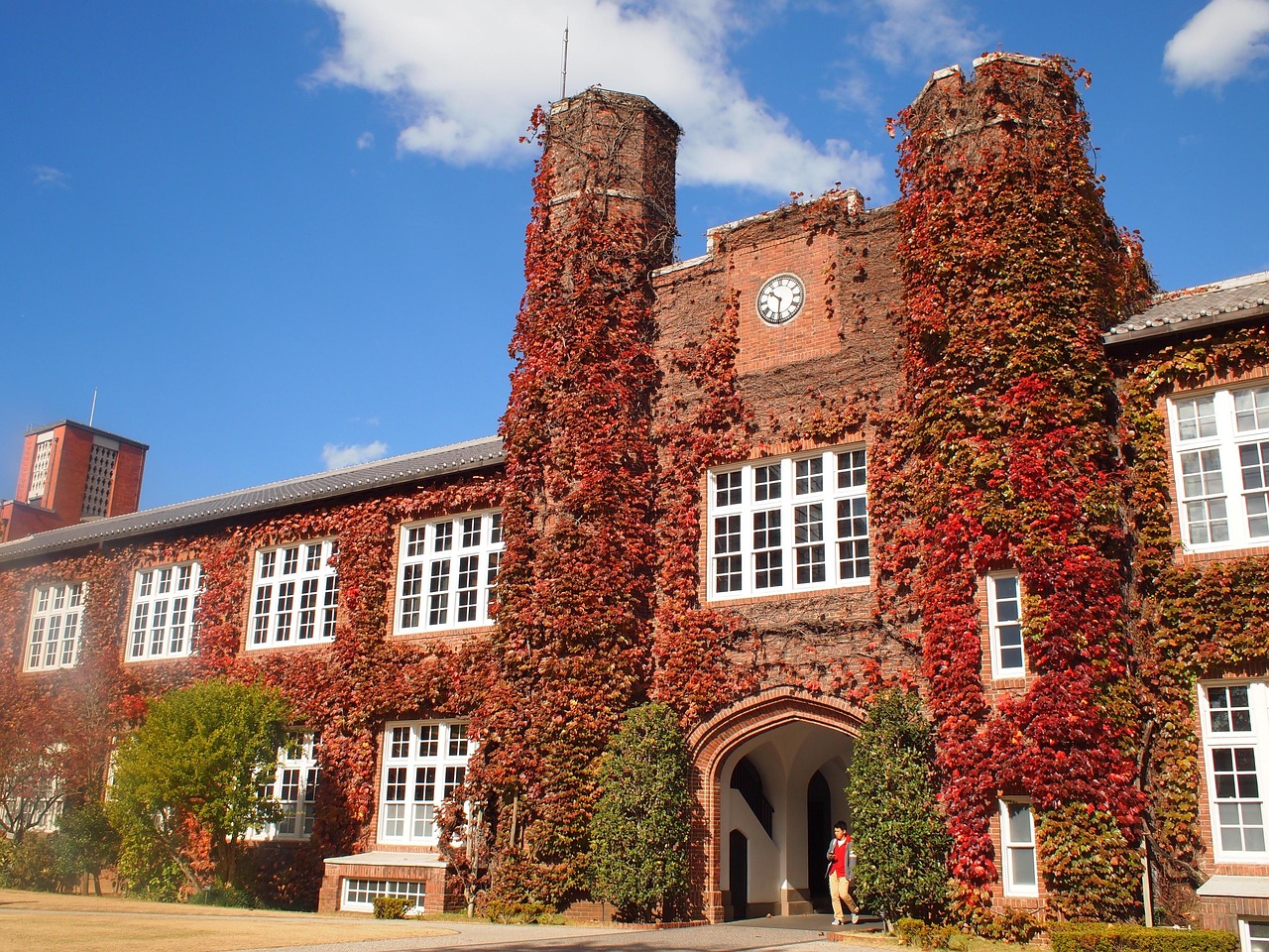 A red-brick university building with two towers and a clock, its walls covered in red ivy under a blue sky