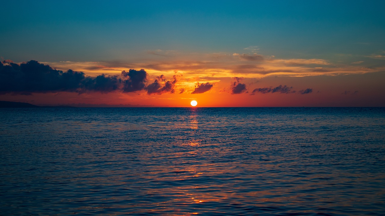 The sun just above the horizon over a calm sea, with an orange sky and dark cloud