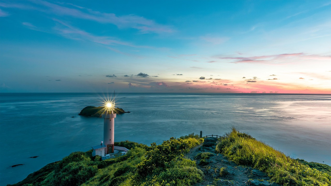 A white lighthouse with its lamp lit on a grassy headland above a calm sea at dusk, a small rocky islet just offshore