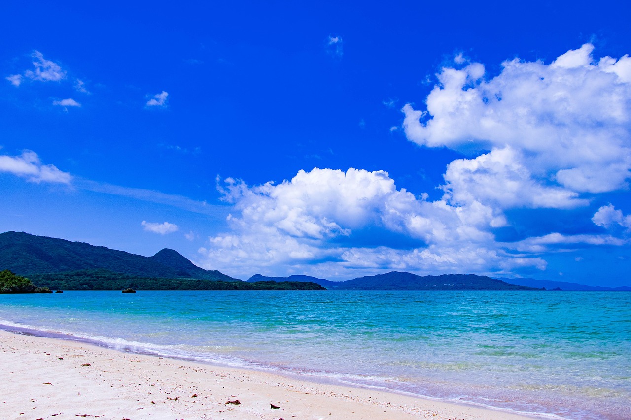 A white sand beach and turquoise shallows under a deep blue sky, forested hills across the water