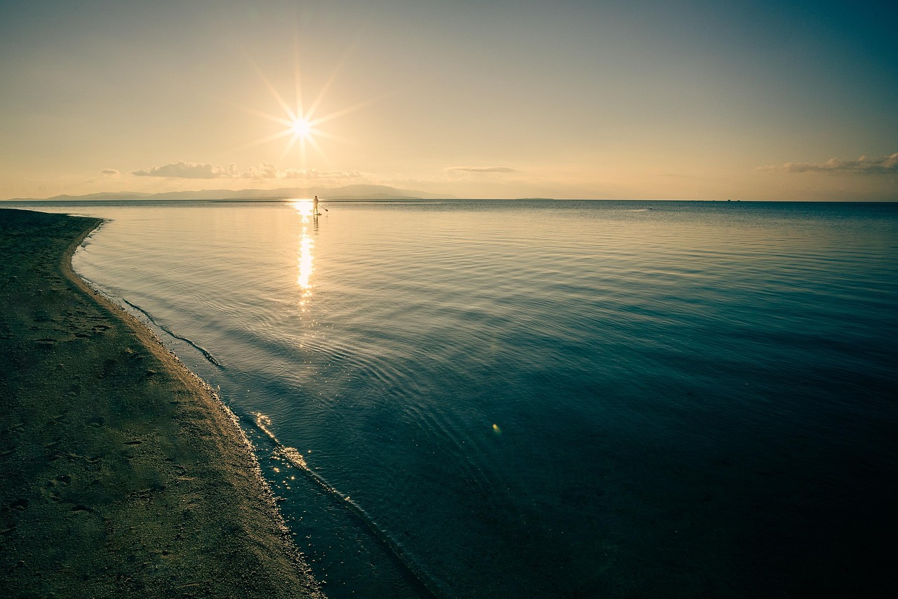 A sandy shore and calm sea at sunset, a distant figure with a paddle on the water