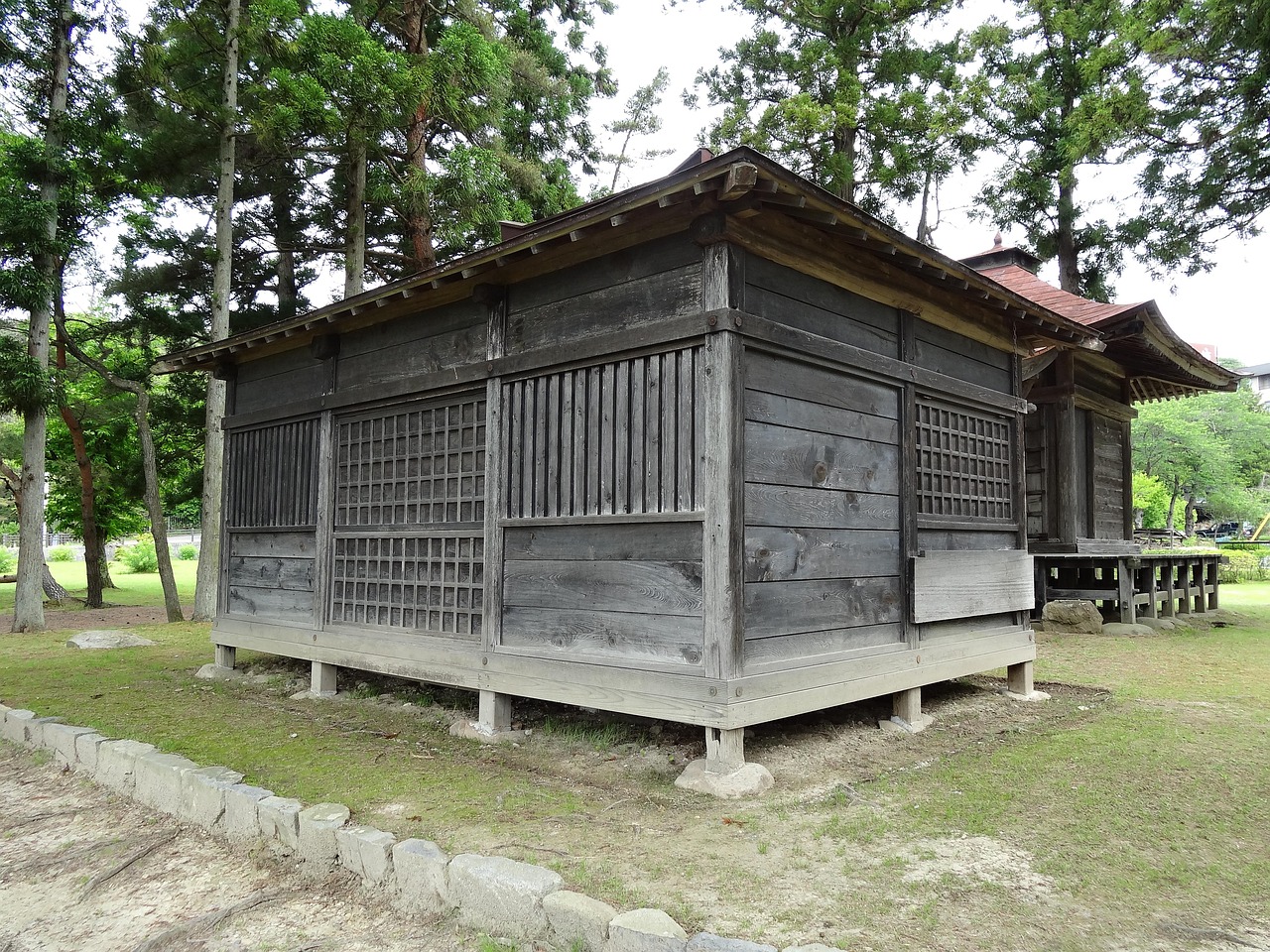 A weathered wooden hall with lattice panels among tall pines on a lawn
