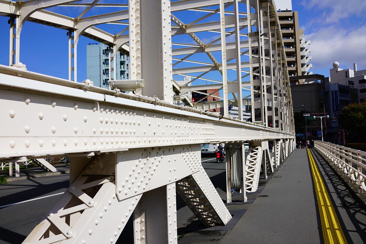 A white riveted iron bridge with a footpath along it, city buildings and blue sky beyond