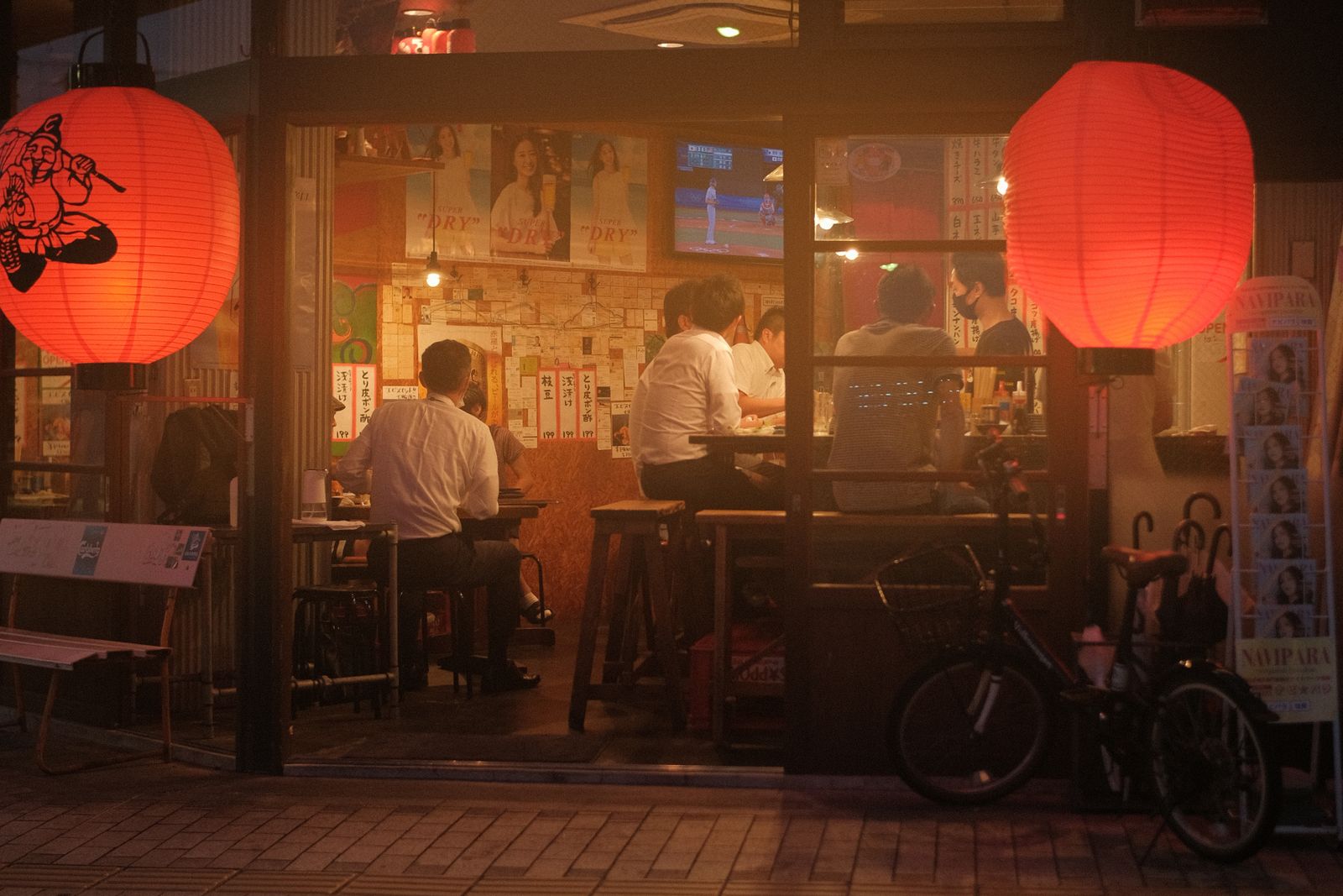 Diners inside a small izakaya, seen from the street through its open front on an evening in Japan.