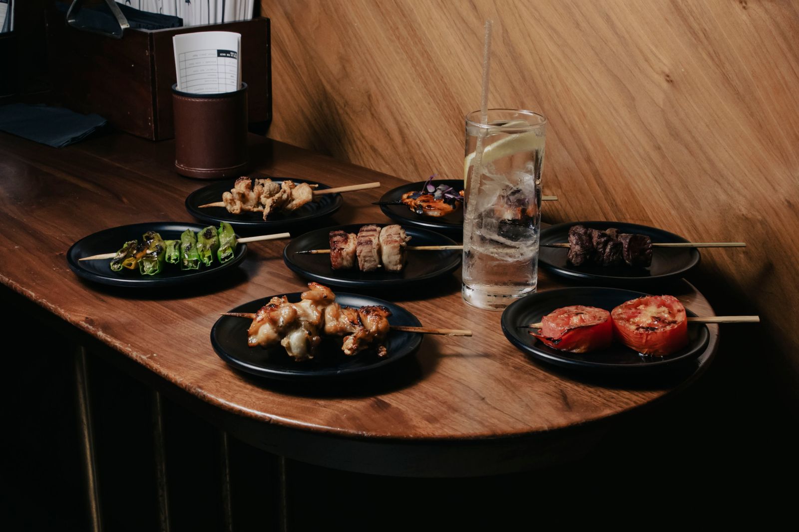 Seven small plates, each holding one grilled skewer, arranged around a tall highball on a wooden counter, with order slips in a leather cup.