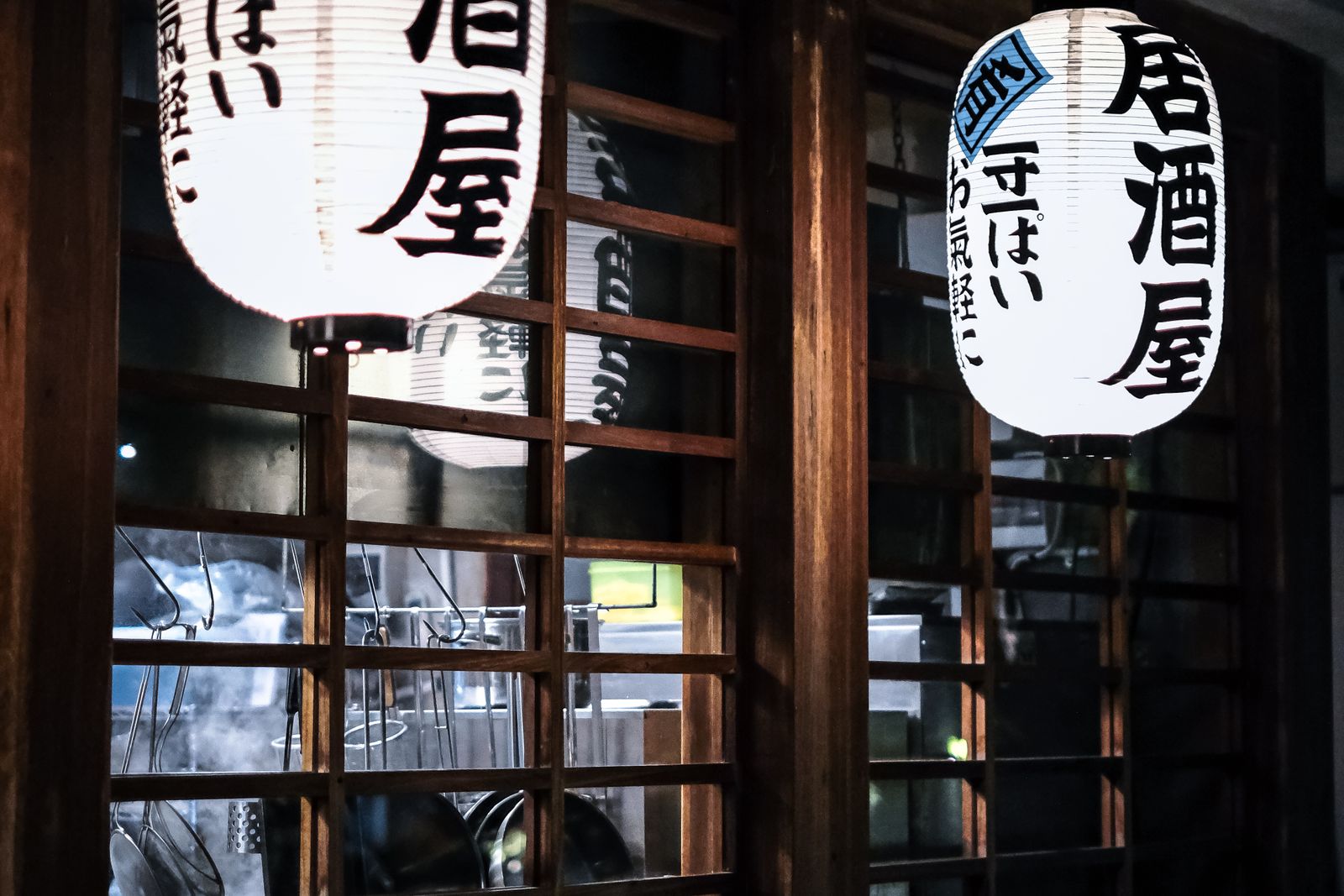 Two white paper lanterns printed with the word izakaya, lit outside a wooden lattice window at night.