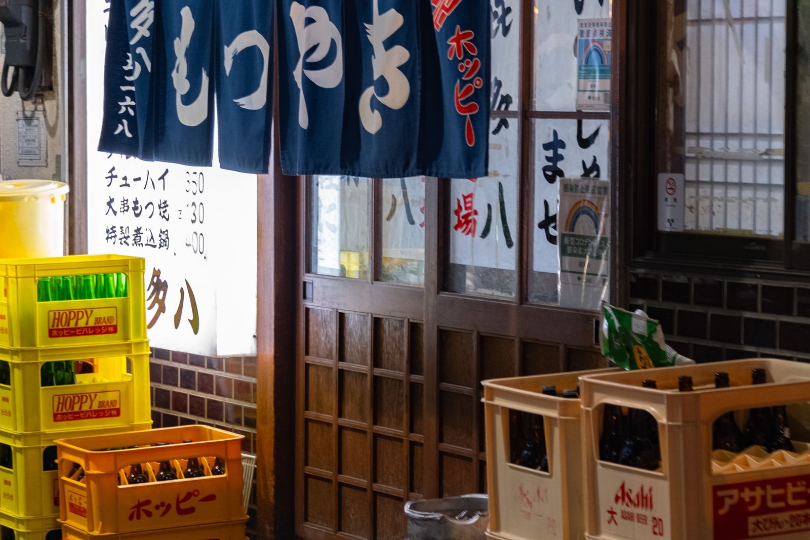 A blue noren curtain hanging over the wooden sliding door of a small Tokyo motsuyaki bar, with bottle crates stacked outside.