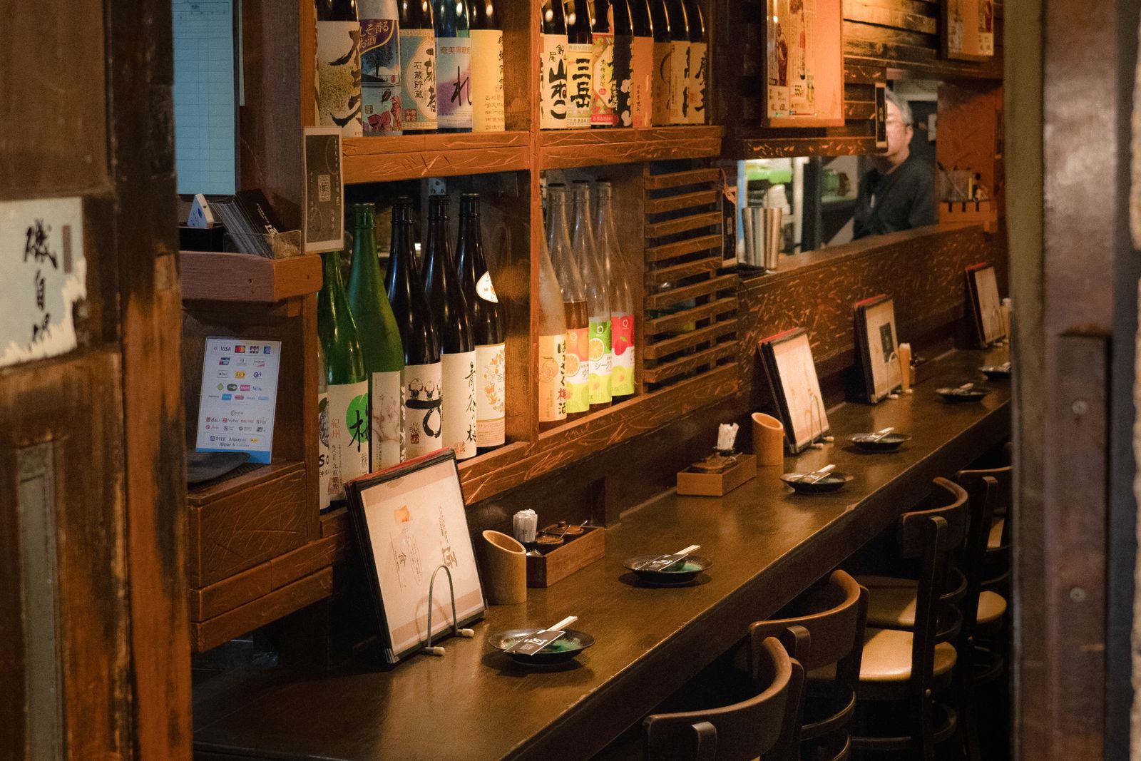 An empty wooden counter set with small plates and menu stands, with shelves of sake and shochu bottles behind it, in Japan.
