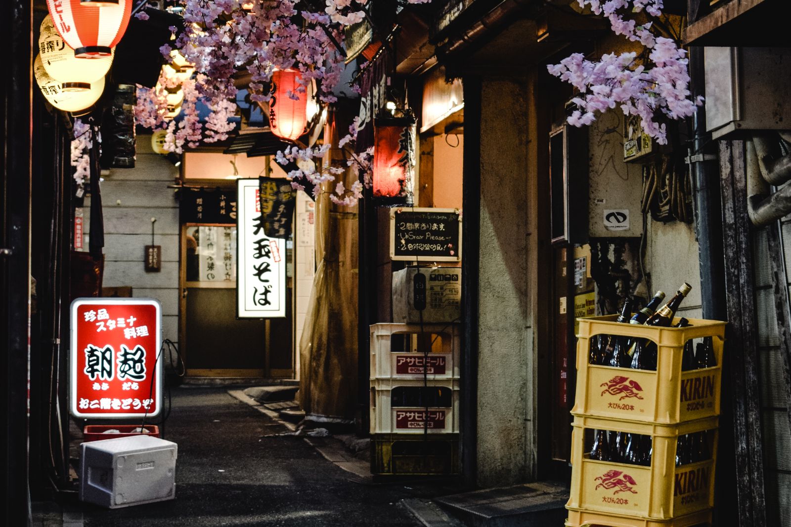 A narrow alley lined with small izakaya at night in Shinjuku, Tokyo, lanterns lit and beer crates stacked along the wall.