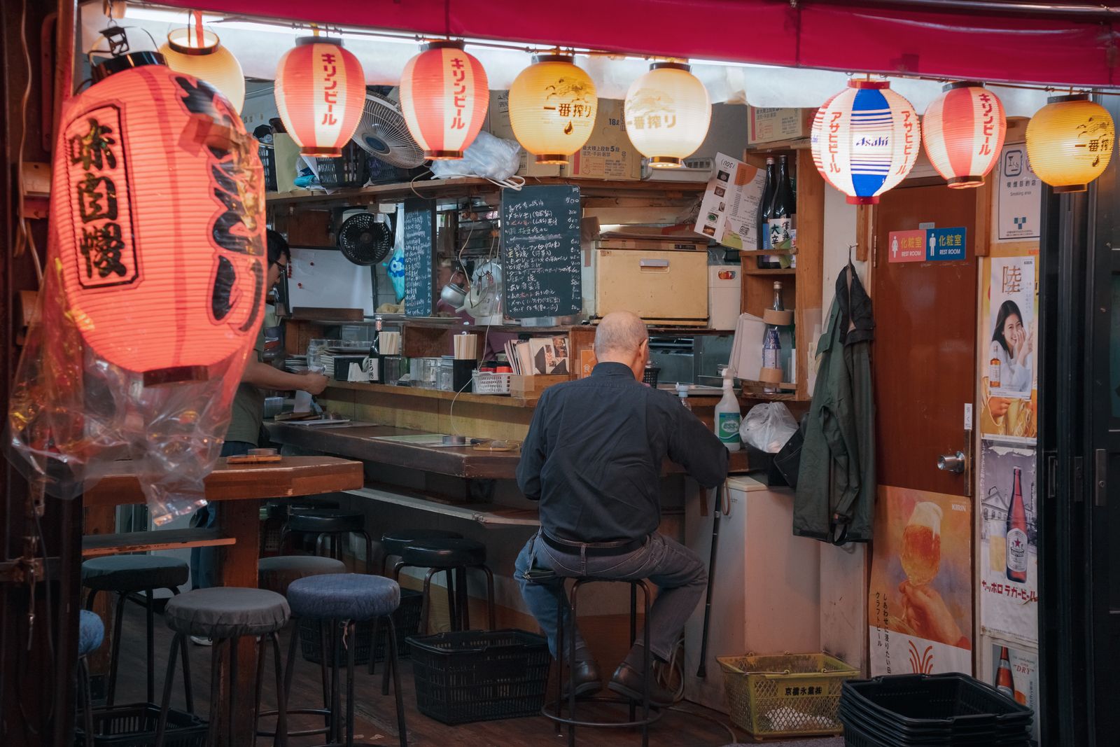 A customer sitting alone at a small izakaya counter in Tokyo, back to the camera, with lanterns and posters lit around the bar.