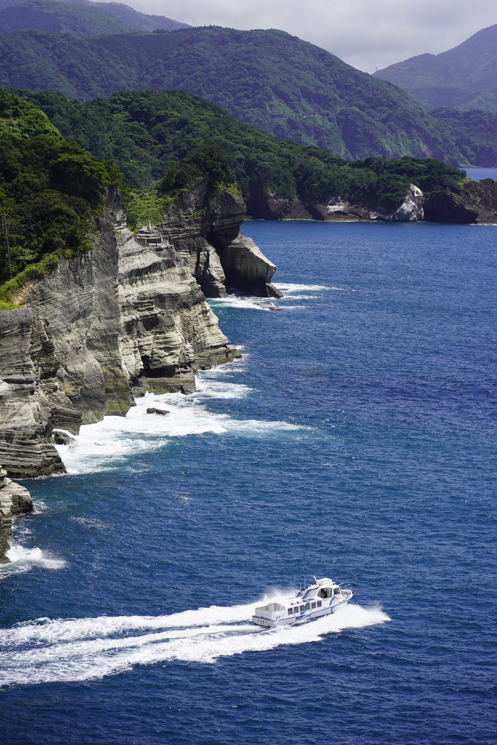 A sightseeing boat passing pale layered sea cliffs on the west coast of the Izu Peninsula, as the photographer places it