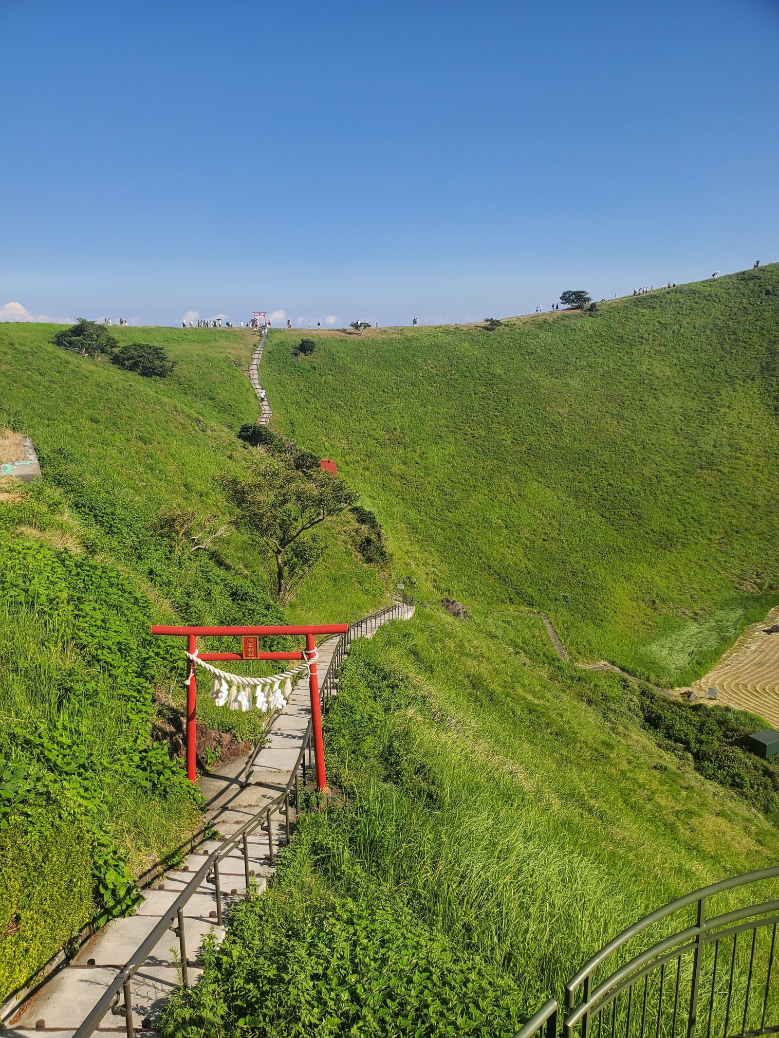 The grassy crater of Mt. Omuro, a red torii on the stair path and small figures walking the rim